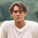 A man sits on a bench with his hands in his pockets, looking contemplative in a serene outdoor setting.