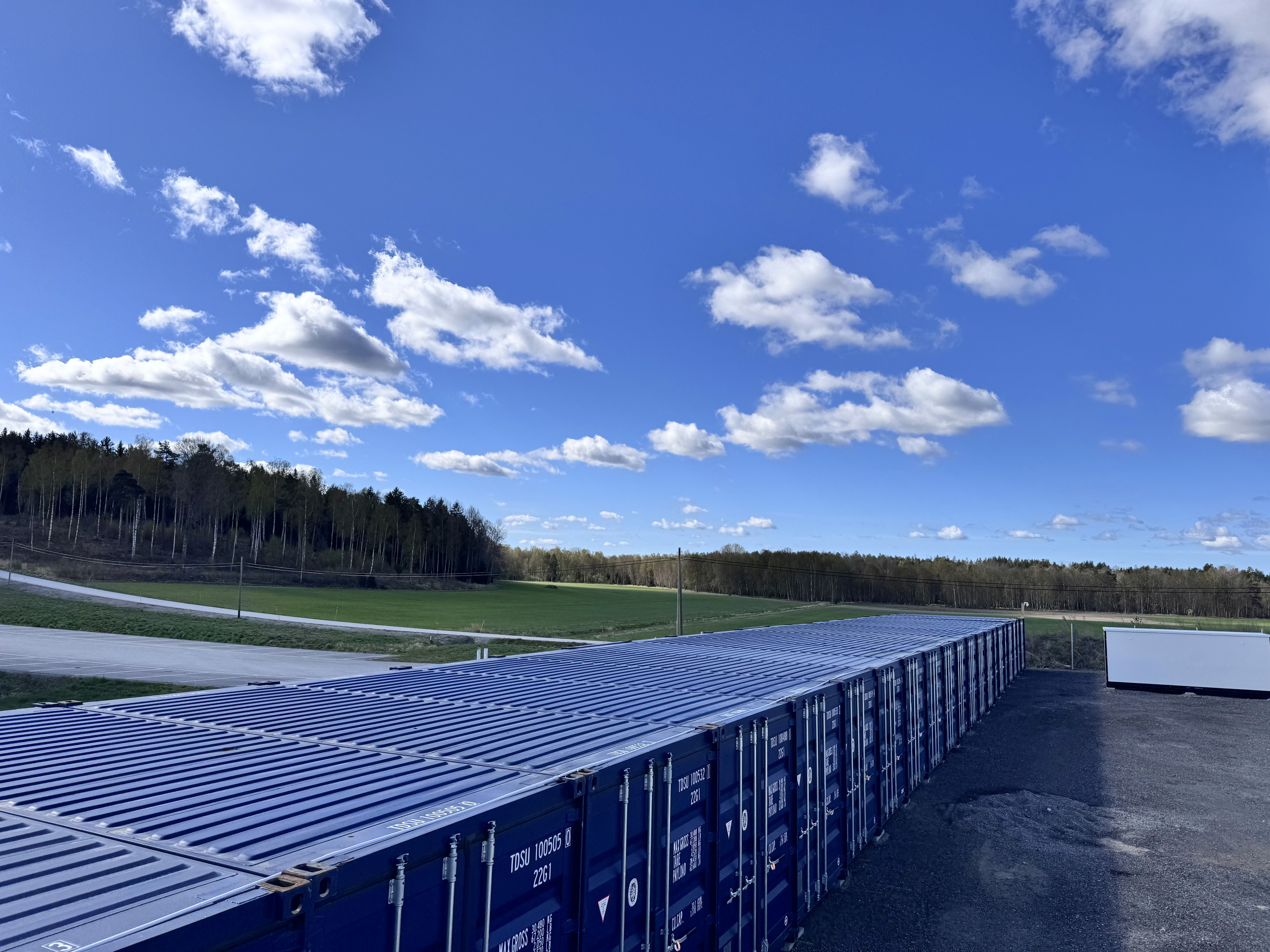 Lång rad blå containerförråd vid öppet fält och skog under klarblå himmel