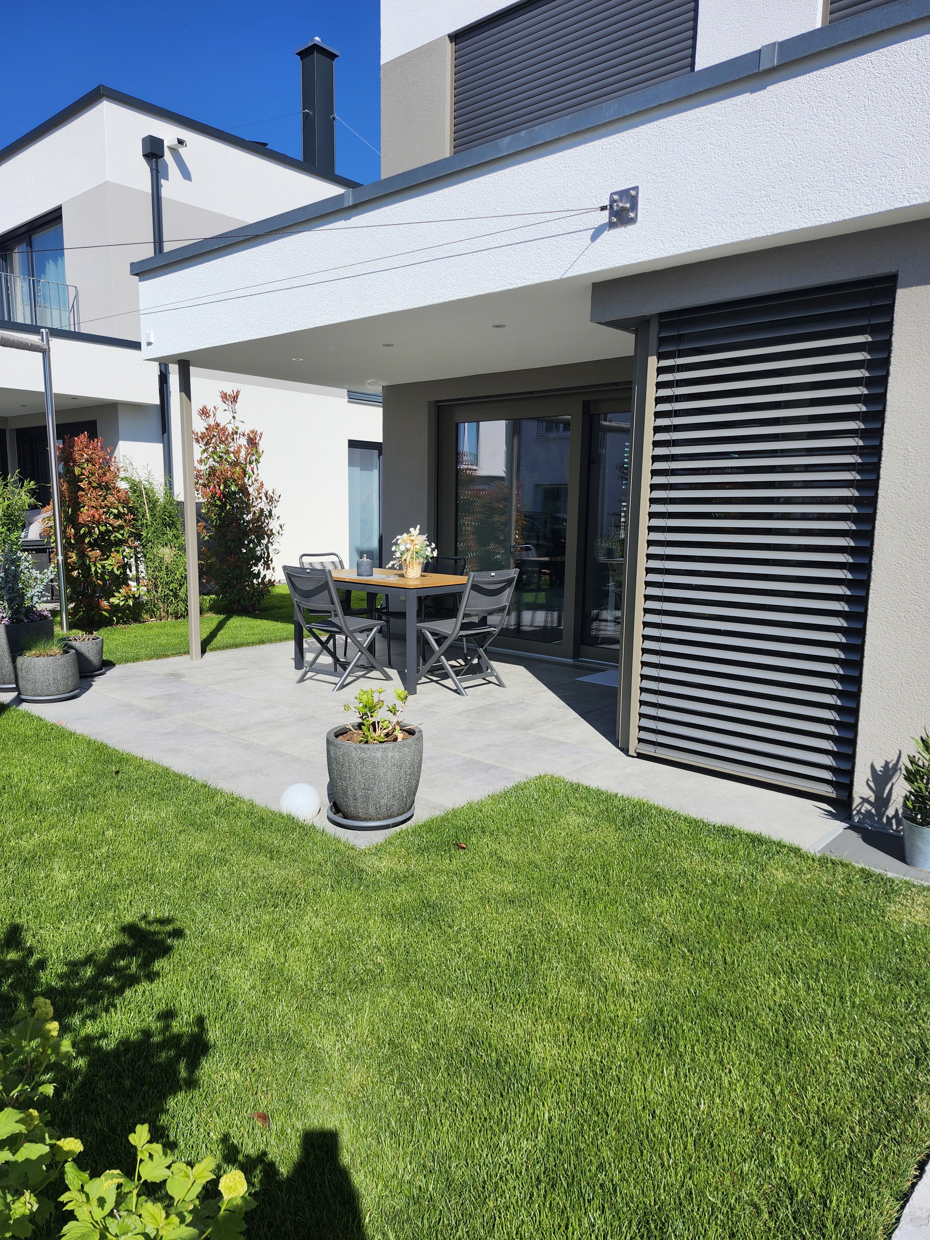 Terrasse mit Esstisch, Stühlen, Pflanzenkübeln und Rasenfläche. Moderne Architektur mit grauen Jalousien. Gartenbau von Marohn & Binder. Sonniger Tag.