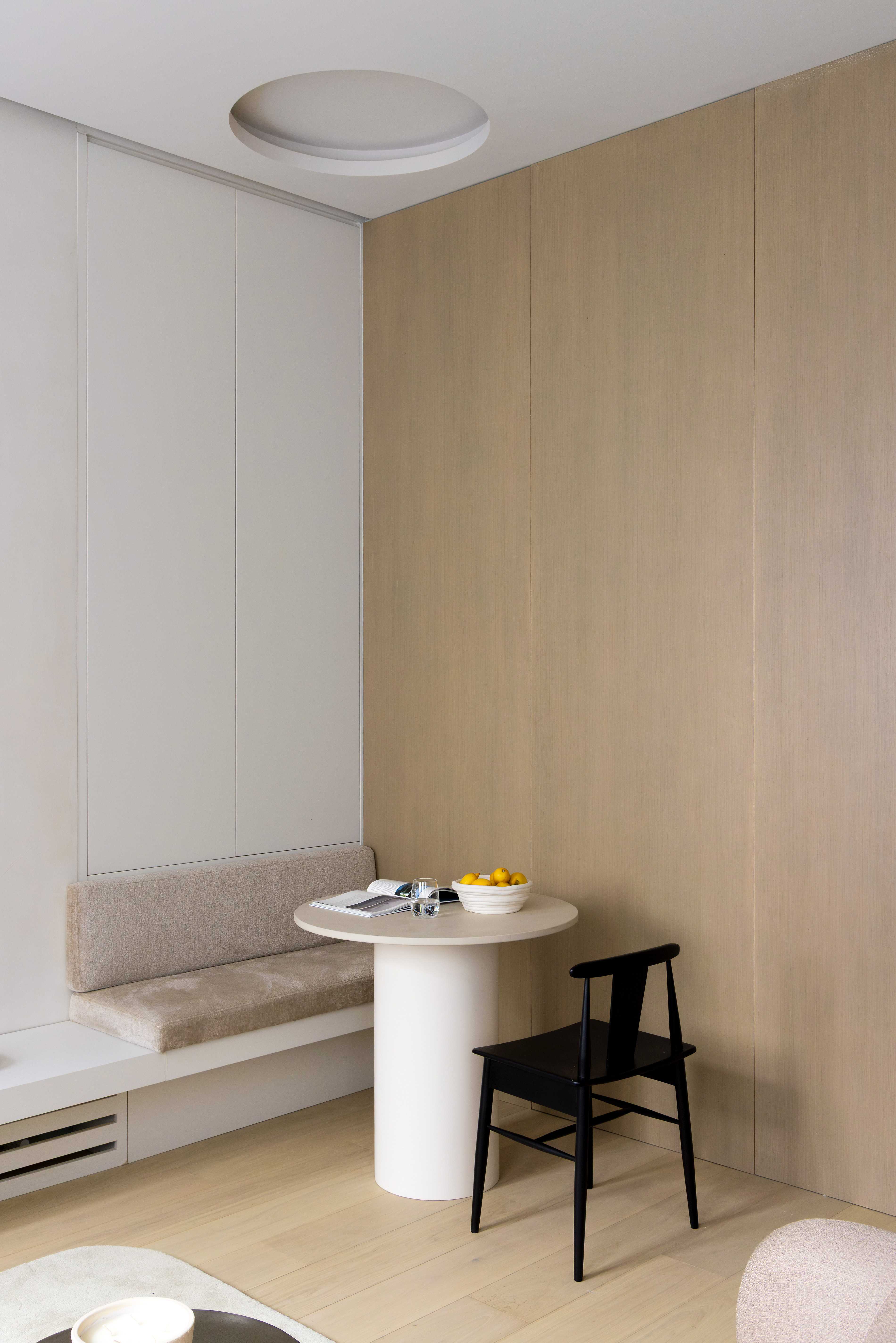 Coin repas vu de face avec banquette beige, table cylindrique blanche, chaise noire et plafonnier encastre