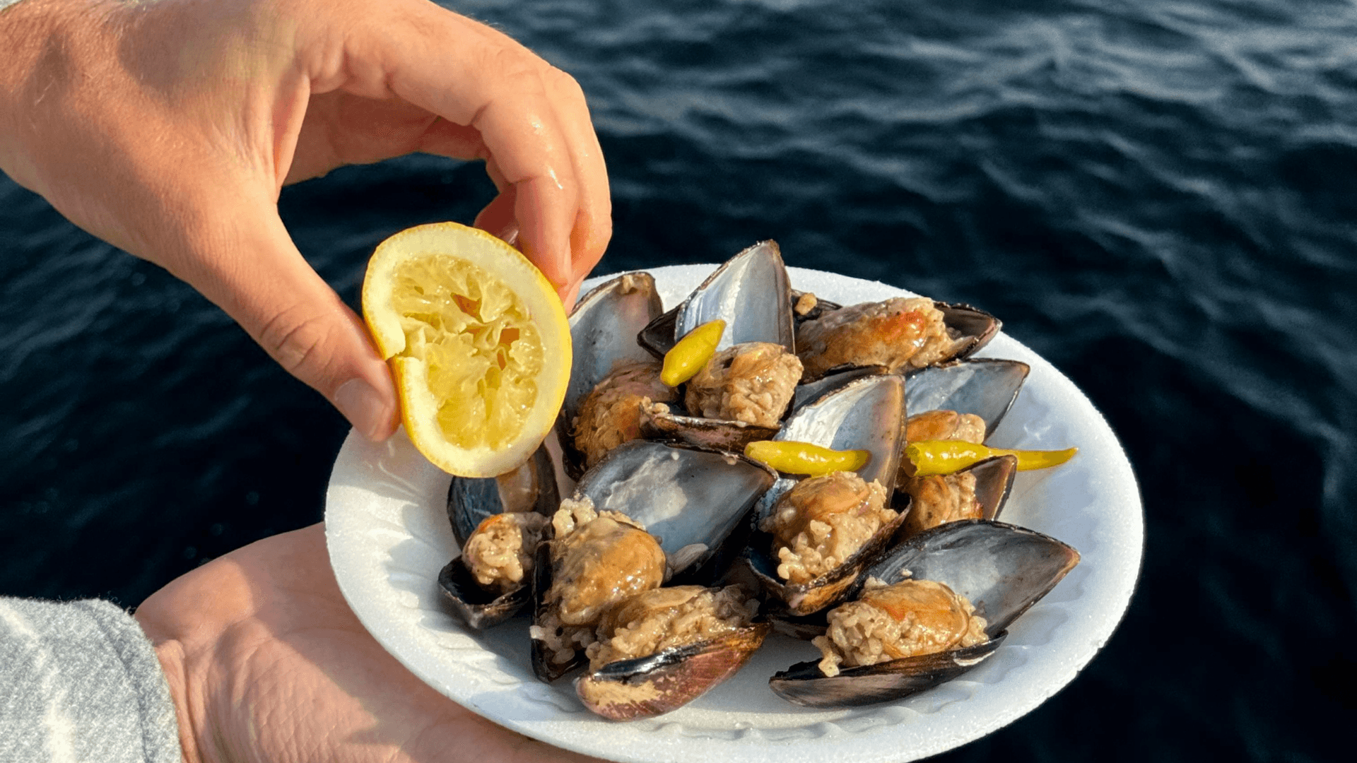 A hand squeezing fresh lemon over a plate of midye dolma — Turkish stuffed mussels with spiced rice — enjoyed by the sea.