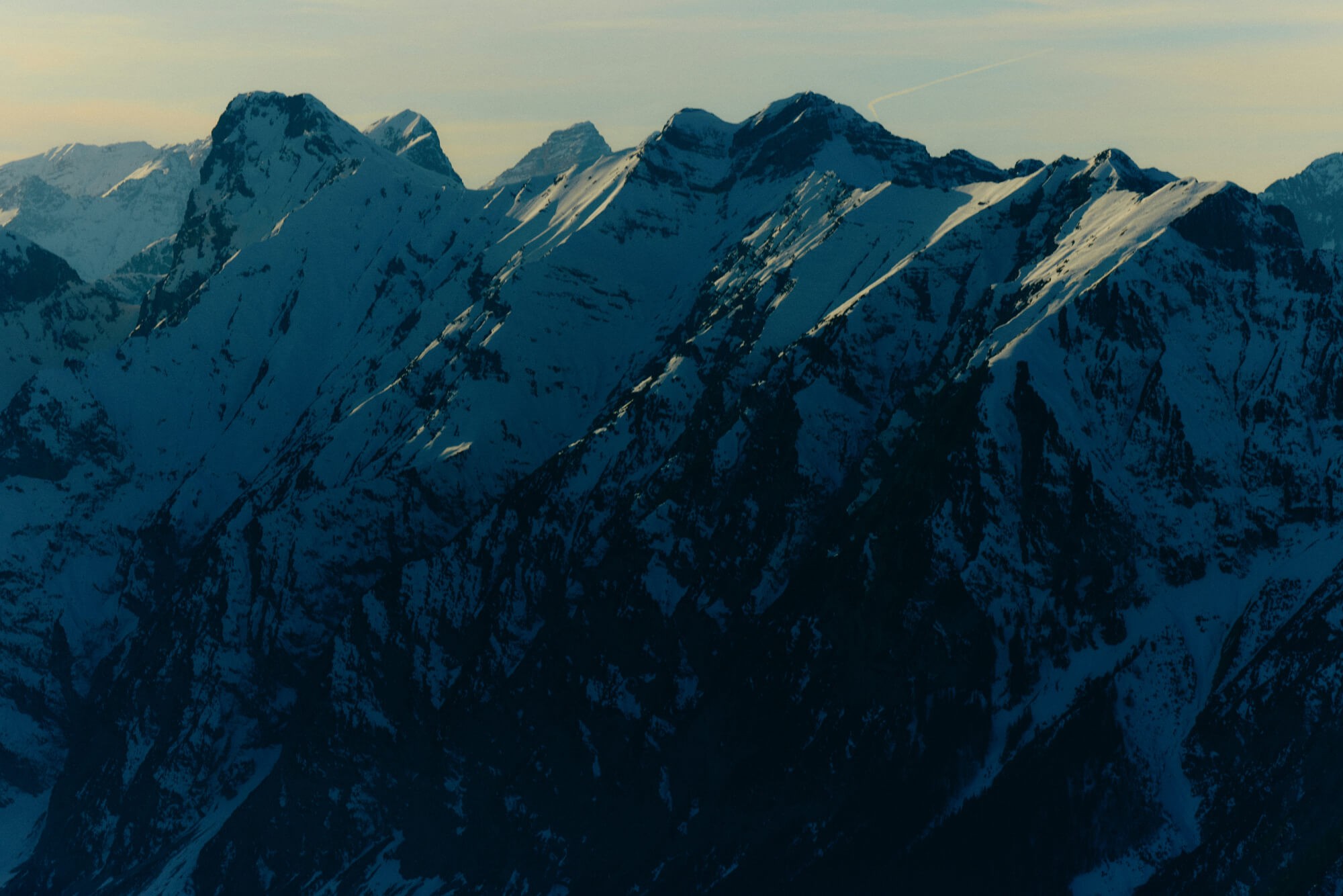 Snow-covered mountain peaks cast in shadow, illuminated by soft sunlight. The sky is clear, evoking a serene and majestic atmosphere.