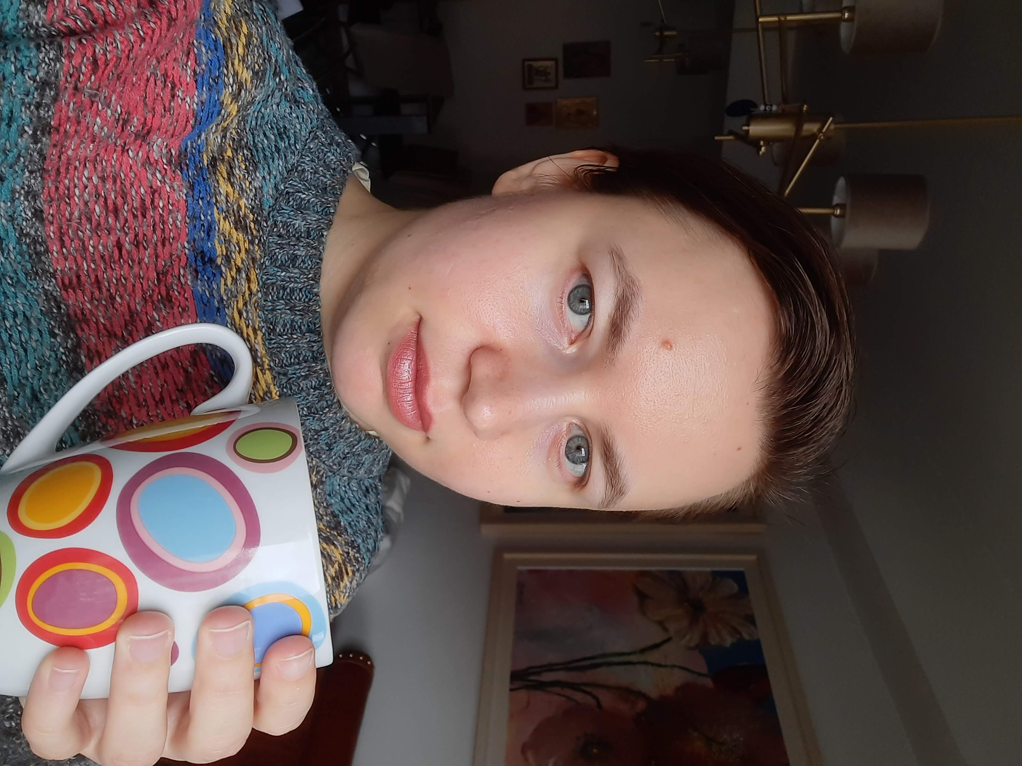 Polina looks at the camera intently, wearing a coloringful sweater and holding a mug of steaming tea.