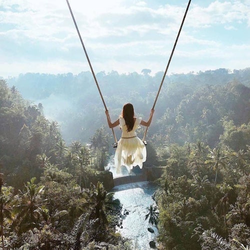 一位穿著白色裙子的女性在明亮多雲的天空下,搖擺在下方有河流的鬱鬱蔥蔥的熱帶山谷上方。