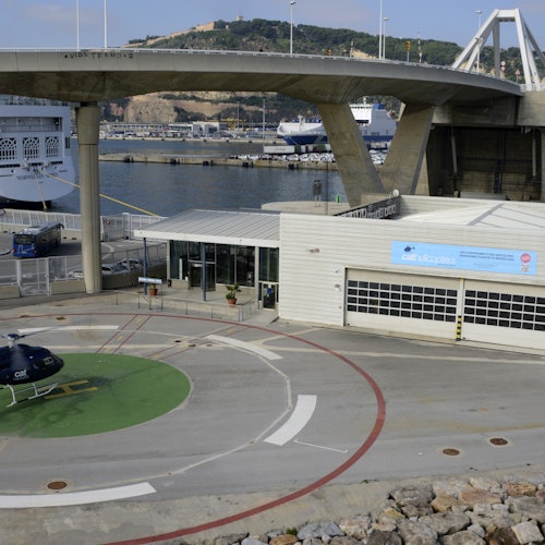 直升机停机坪位于港口一栋建筑旁边,背景是桥梁和一艘大船。