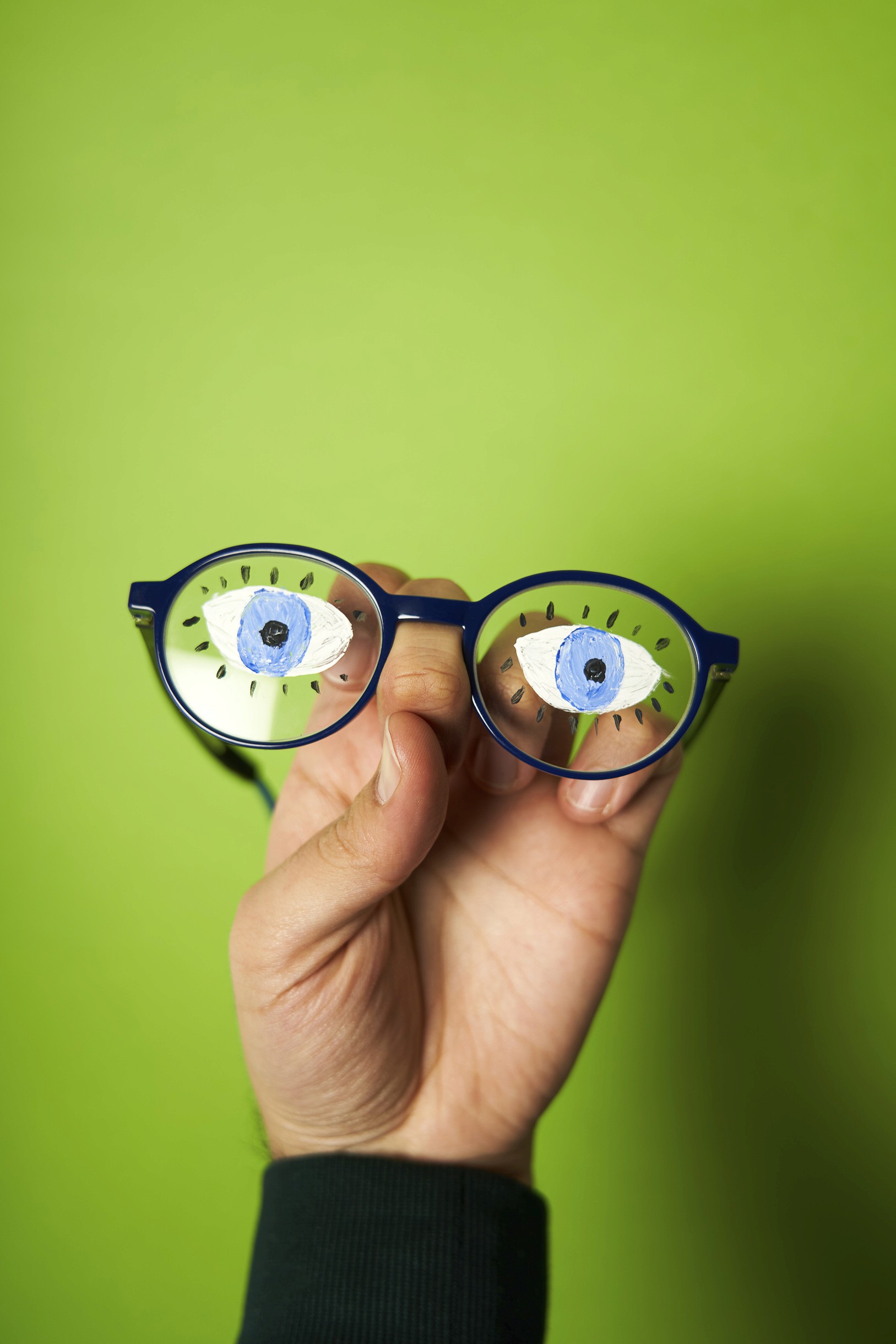 Photographie studio sur un fond vert pomme d'une main portant une paire de lunette de lecture bleue avec des yeux dessinés sur les verres