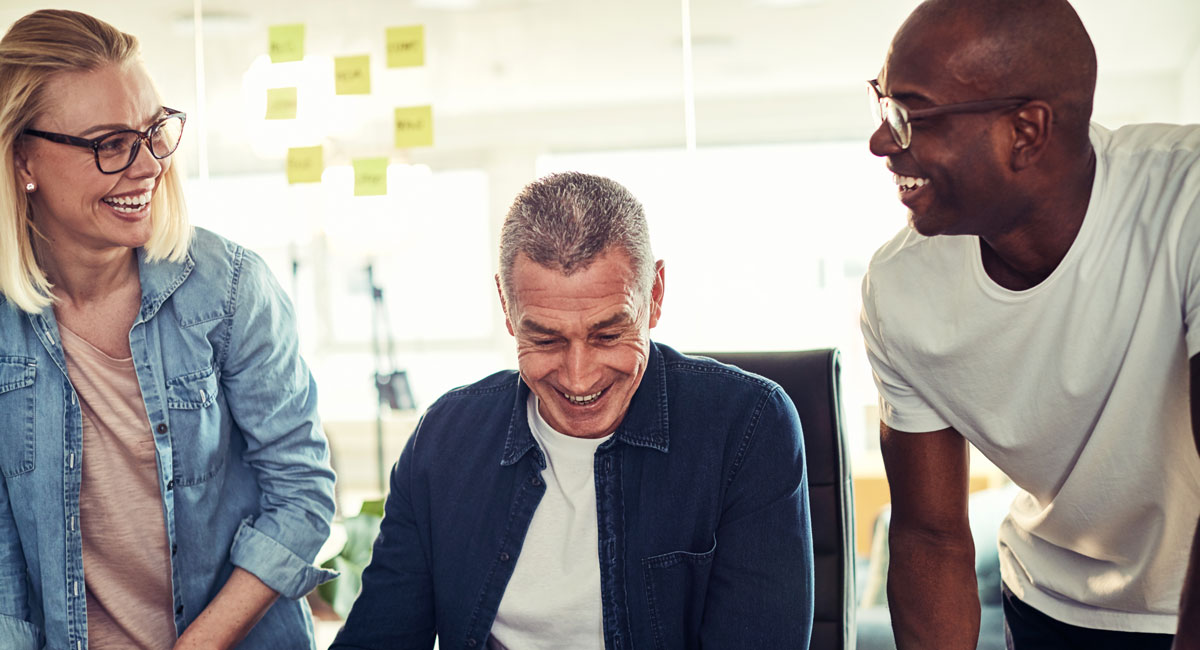 Three diverse coworkers laughing together near a glass board with sticky notes in an office