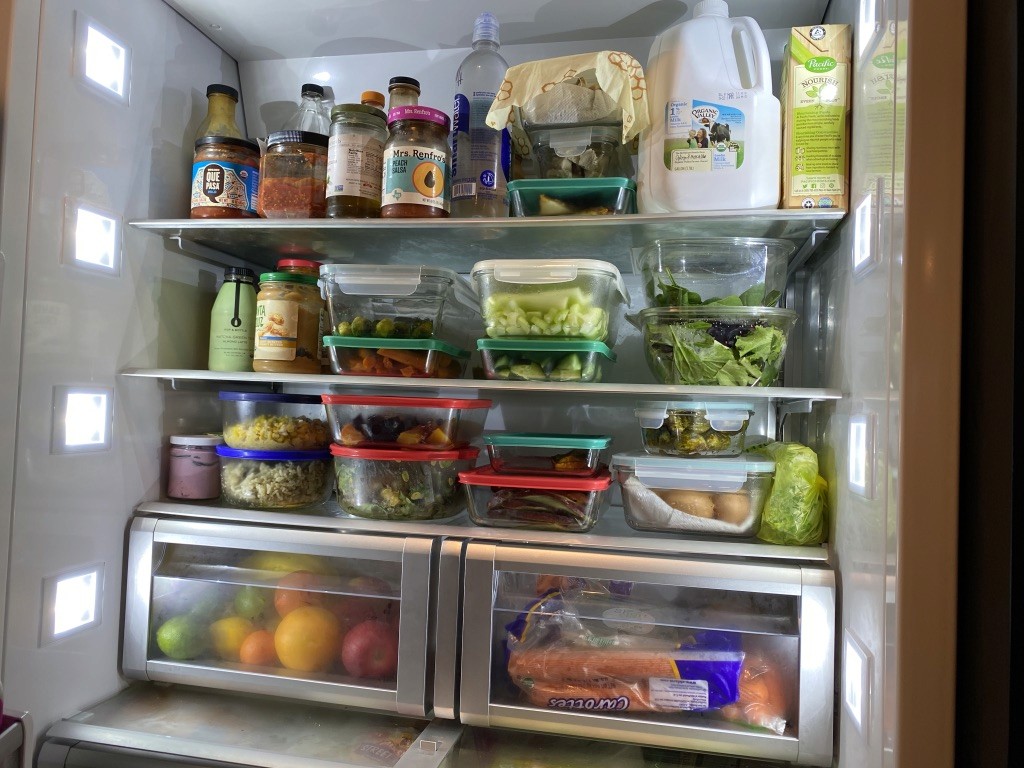 An open refrigerator filled with various items, including containers of fresh vegetables, bottled sauces, packaged juices, neatly stacked leftovers, and a variety of fruits in the drawers below.