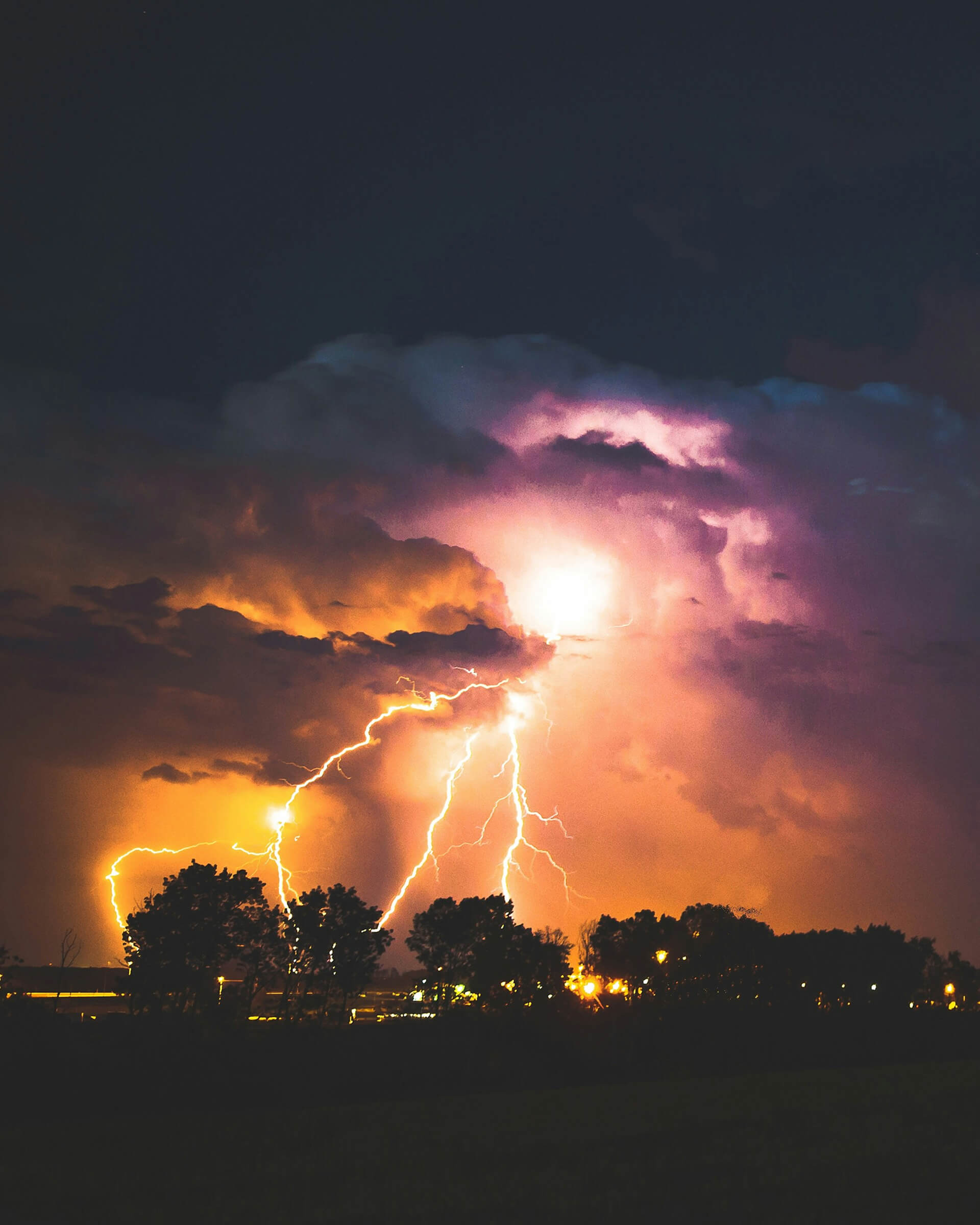 Dramatic photo of a lightning storm at night, with multiple bright bolts striking near a silhouetted treeline. The sky is illuminated in shades of orange, purple, and white, creating a powerful contrast against the dark foreground.