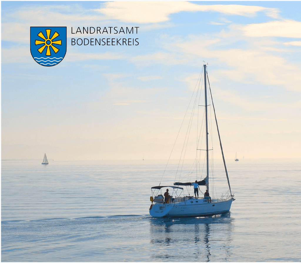 Ein Segelschiff fährt am Morgen auf ruhigem Wasser die Sonne scheint langsam aufzugehen, darauf befindet sich das Logo des Landratsamt Bodenseekreis