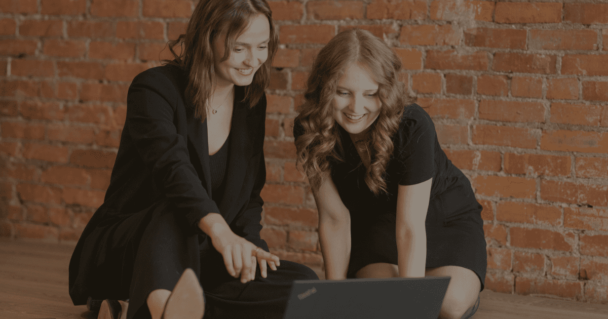 Two fair skinned women kneeling on a floor looking at a laptop. They are wearing black dress attire. In the background is red brick and the floor is hardwood.