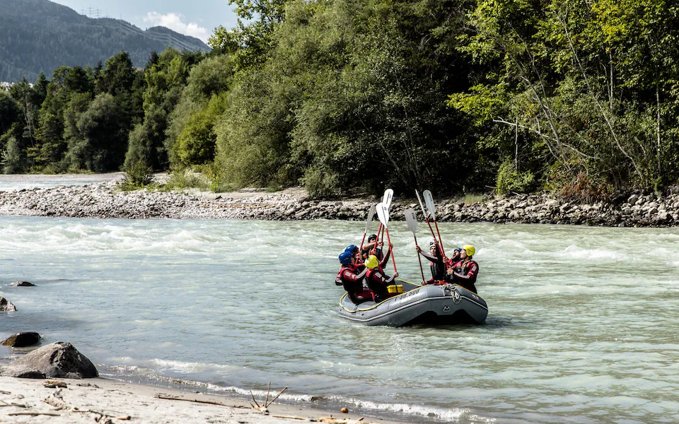 Group of people white water rafting on a scenic river surrounded by forested mountains, raising paddles in celebration during an outdoor adventure.