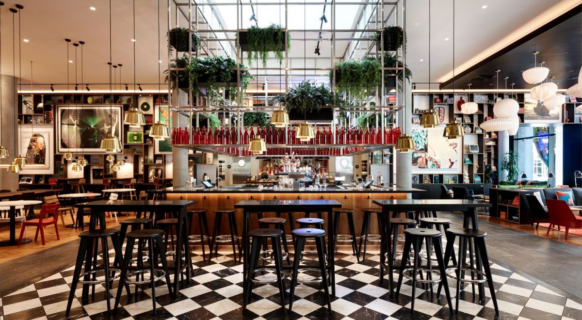 The 'canteenM' hotel bar features dark bistro tables on a checkered floor, a suspended metal bar rack adorned with red bottles, and floating plants.