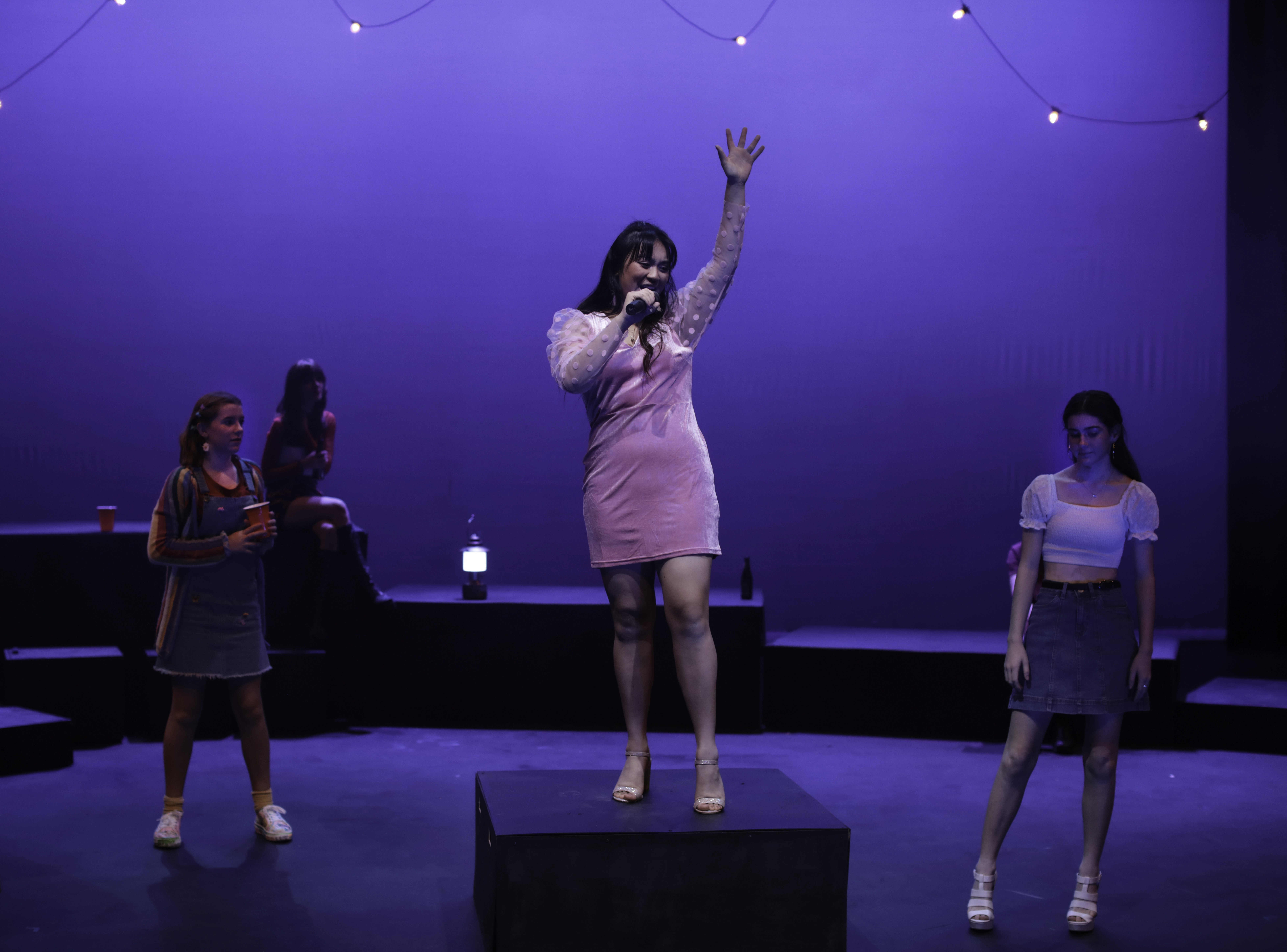 A teenage girl in a pink party dress stands on a platform, singing confidently into a microphone with one arm raised. Other teens watch nearby, surrounded by soft string lights and a blue nighttime atmosphere.