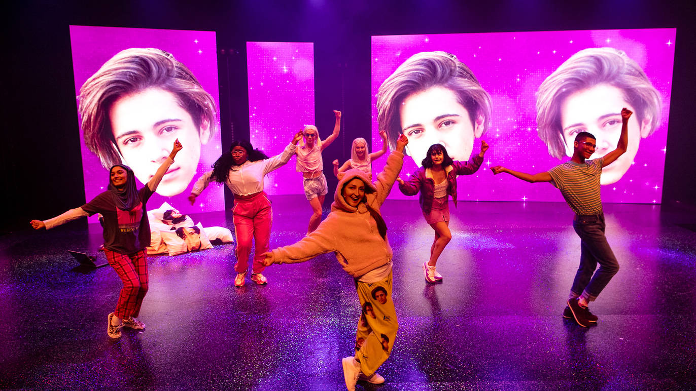 A cast of eight performers dance energetically across a glittering stage in front of two giant pink screens featuring a teen idol’s face.