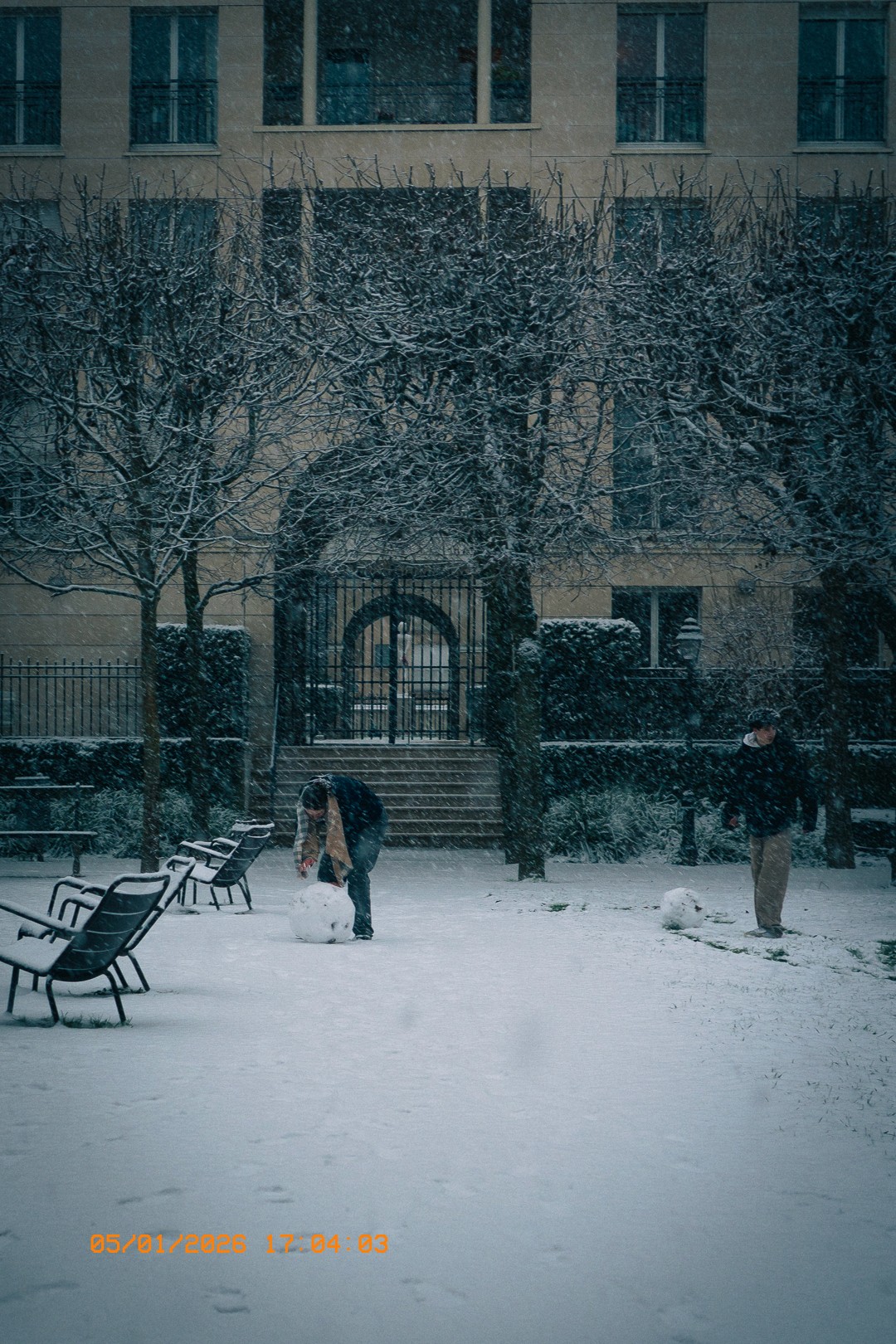 Jardin des Mille Roses de l’Hôpital Corentin Celton sous la neige, avec deux personnes construisant des bonshommes de neige. Des arbres enneigés, des bancs et une pluie de flocons.