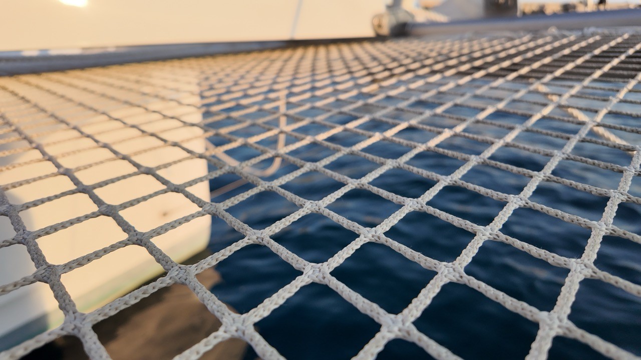 Close-up of the woven white trampoline netting stretched between the bows of a Lagoon 42 catamaran, with blue water visible below and warm evening light on one hull.