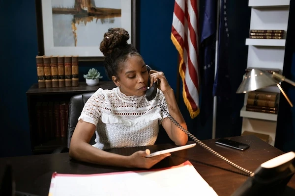 A woman in a white blouse sits at a desk in an office with an American flag, talking on the phone while holding a tablet.