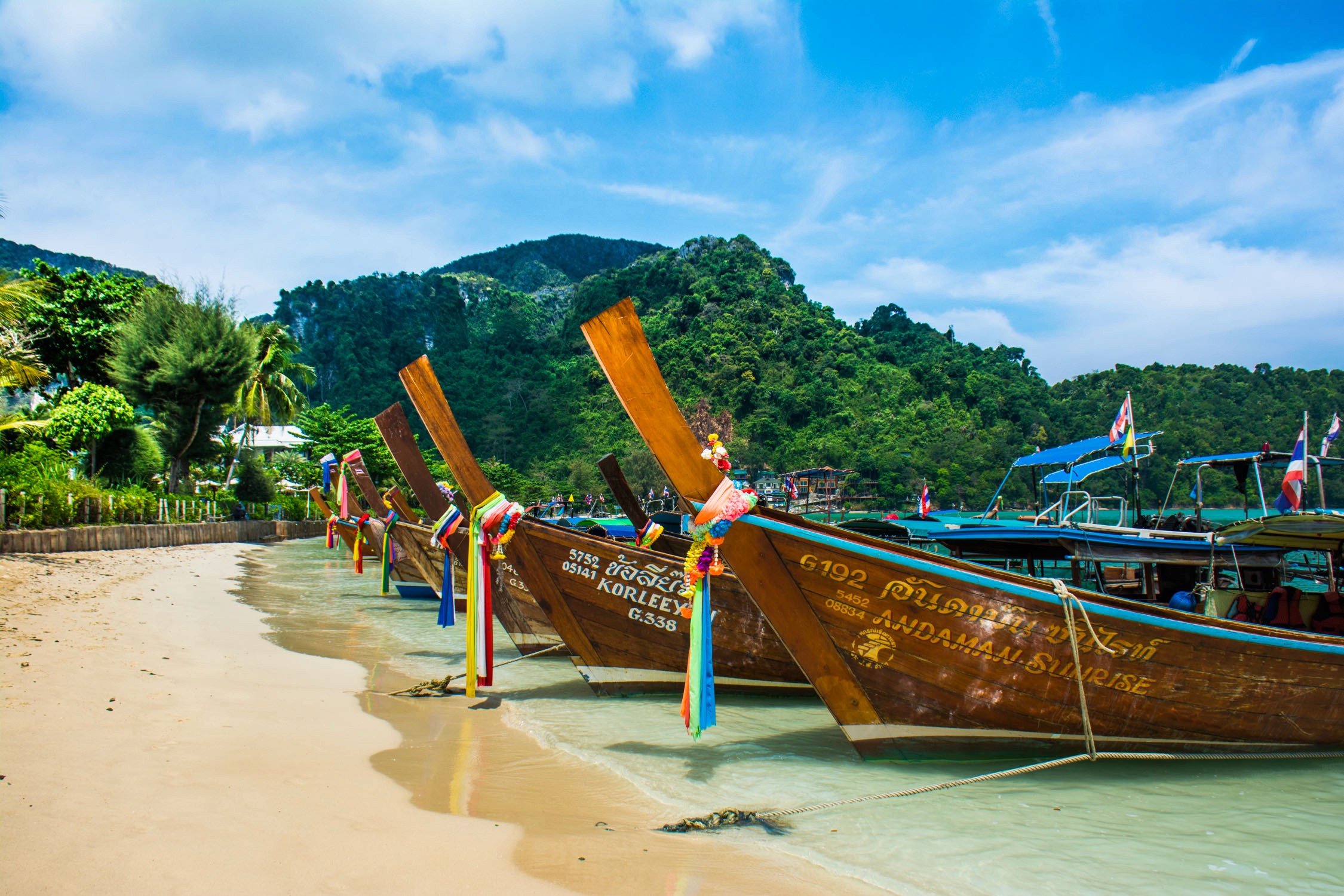 Colorful longtail boats rest on sandy shores under bright blue skies near lush green tropical mountains.