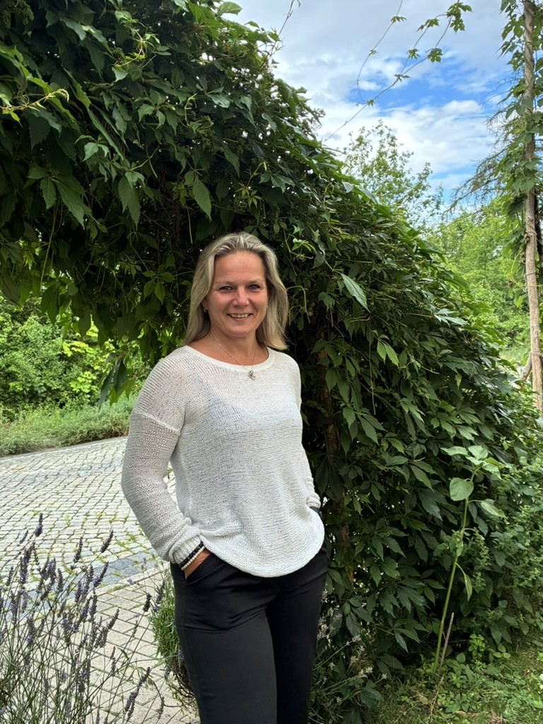 Danica Homann, eine lächelnde Frau steht vor einem schönen grünen Pflanzenbogen. Sie trägt einen weißen Pulli und hat die Hände lässig in die Taschen gesteckt.