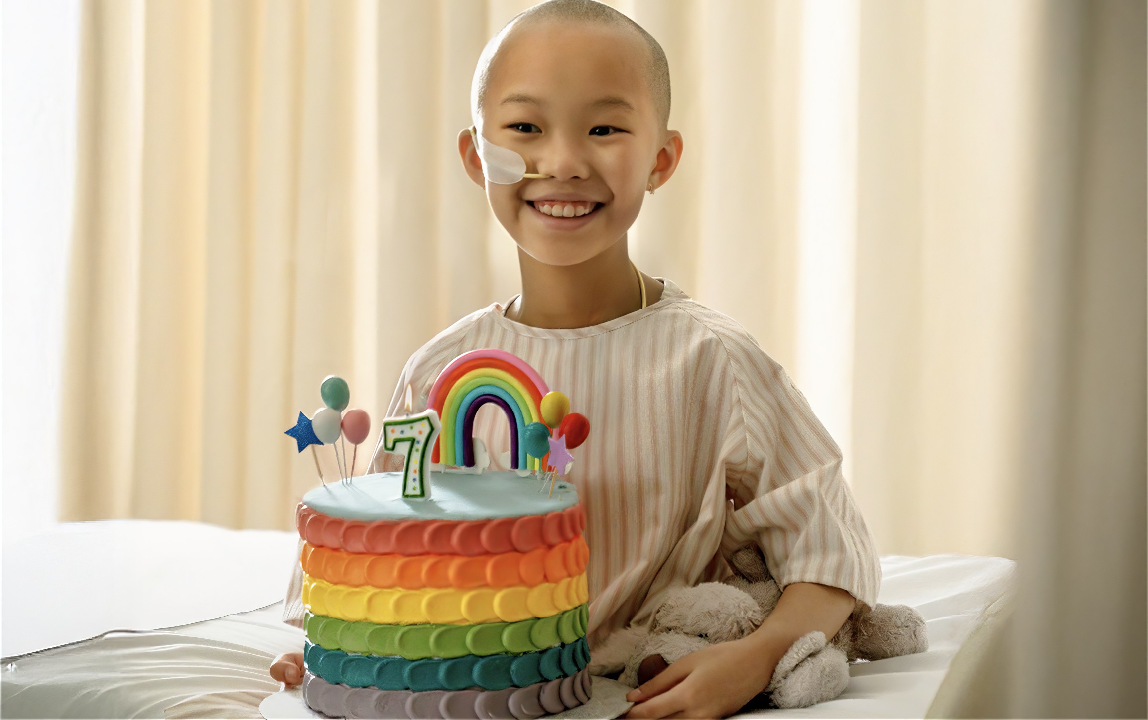 Smiling child in a hospital setting with a colorful rainbow birthday cake and number seven candle.