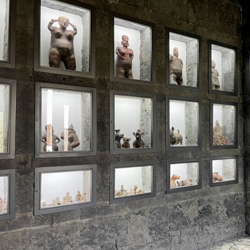 Isang pader na nagpapakita ng maliliit, sinaunang-tulad na mga pigura ng tao sa loob ng mga glass box sa isang istrukturang bato.