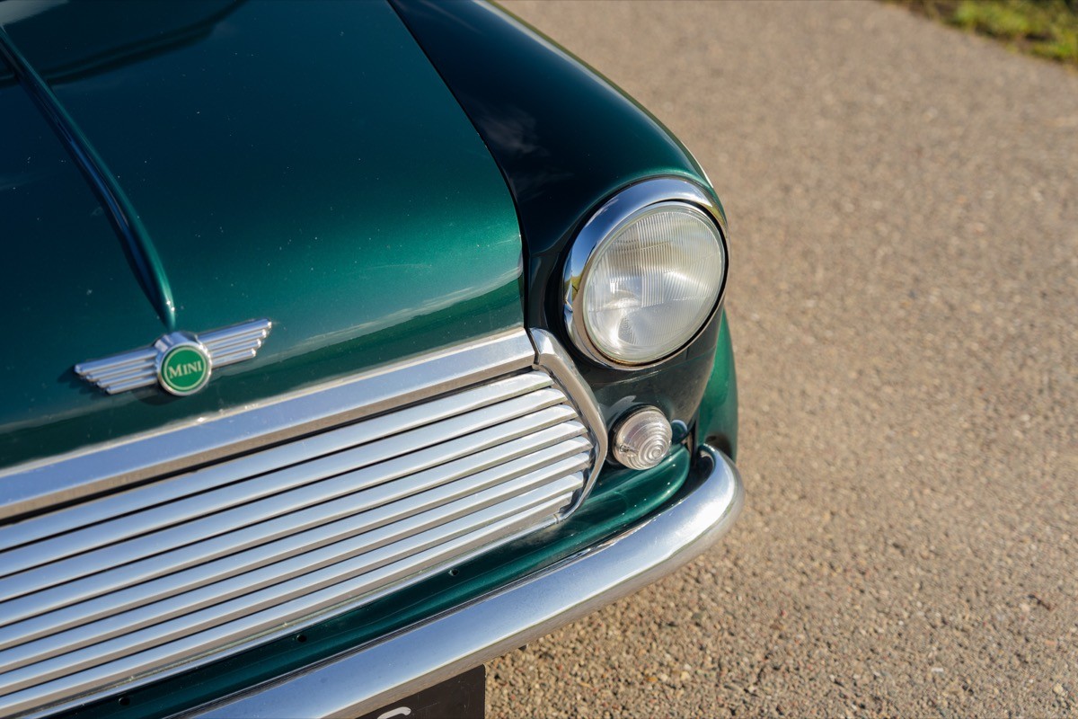 Close-up of a vintage green car's front hood and headlight, parked on a gravel surface.