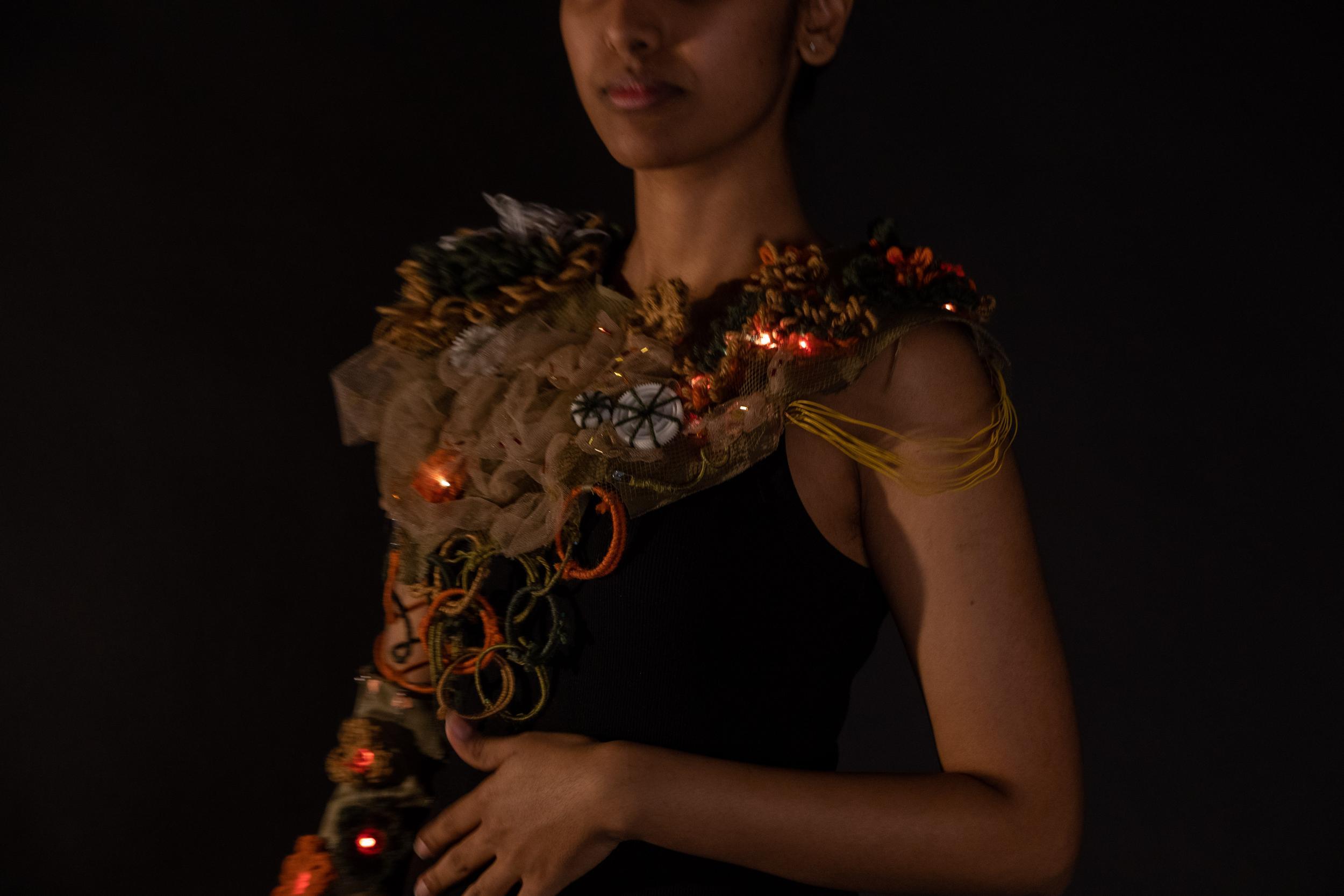 A close-up image of a wearable technology garment. A person's hand reaches out to touch a circular tactile sensor on the shoulder, which triggers a sequence of glowing orange LED lights embedded within the textured, soft-circuitry fabric. The garment features intricate layers of yarn, mesh, and exposed wiring.