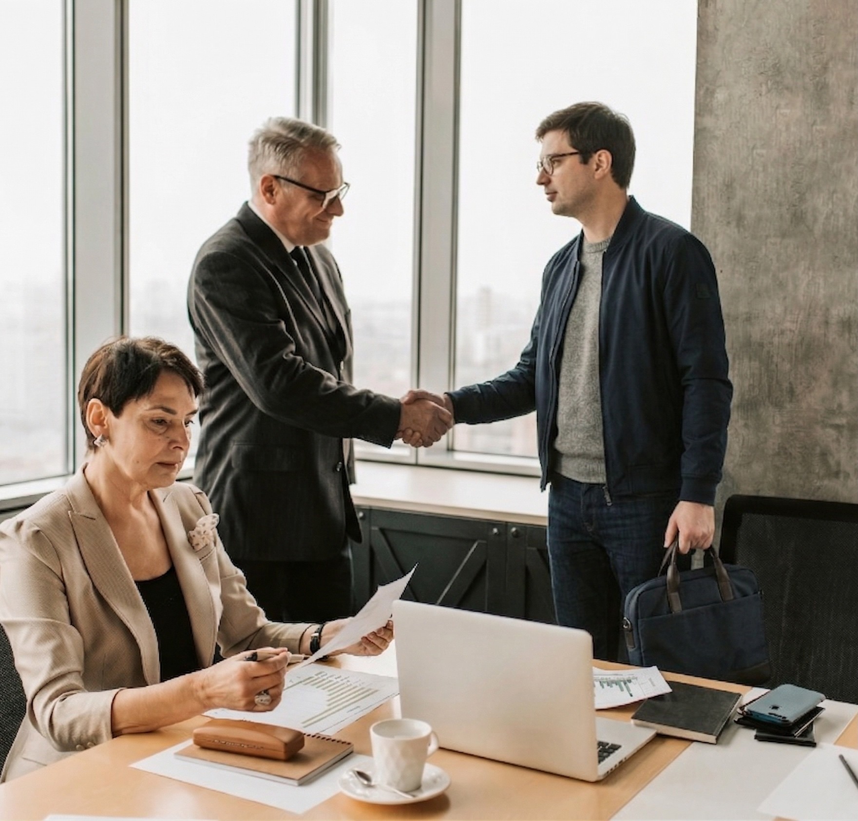 Deux personnes se serrant la main dans une salle de réunion, tandis qu’une collègue consulte des documents à son bureau