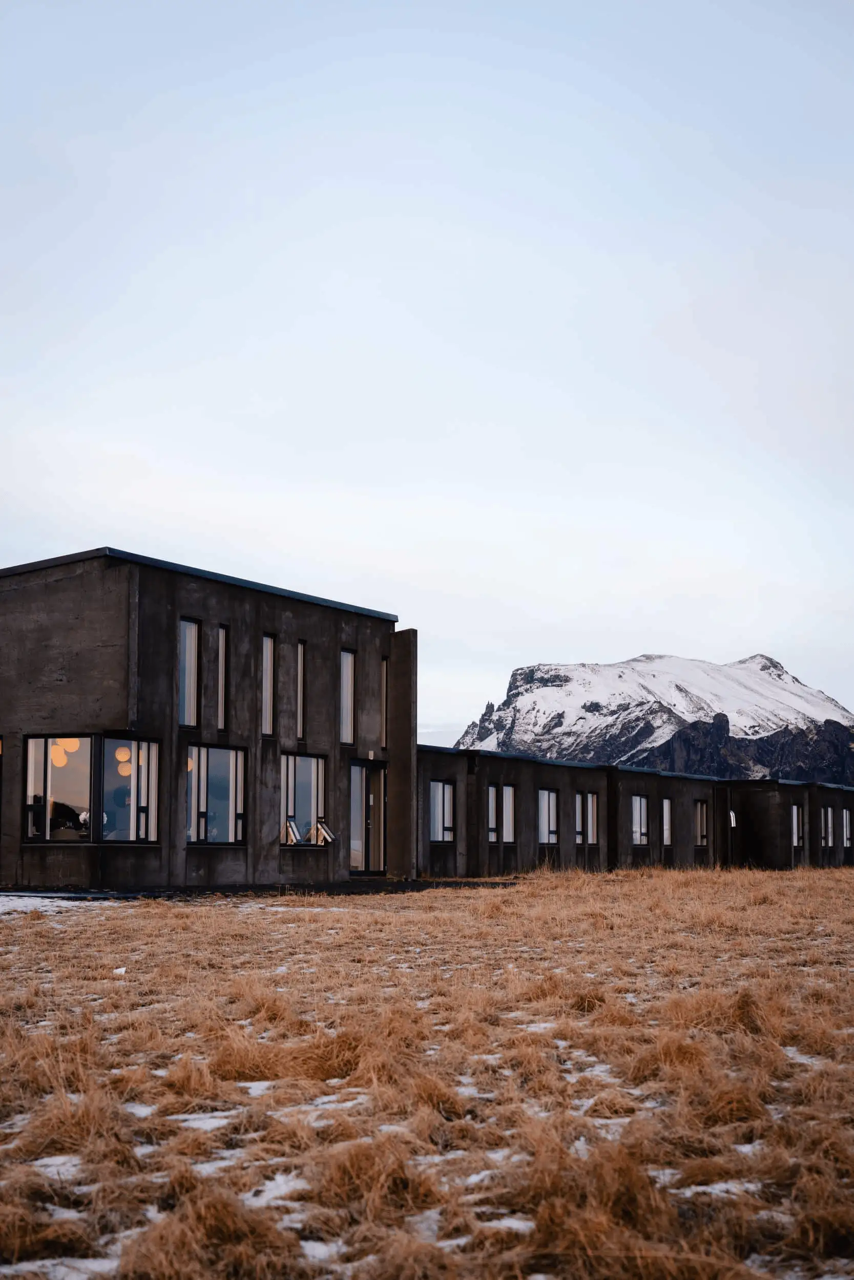 Dark concrete hotel building on a field of dry grass, with a snow-capped mountain behind it