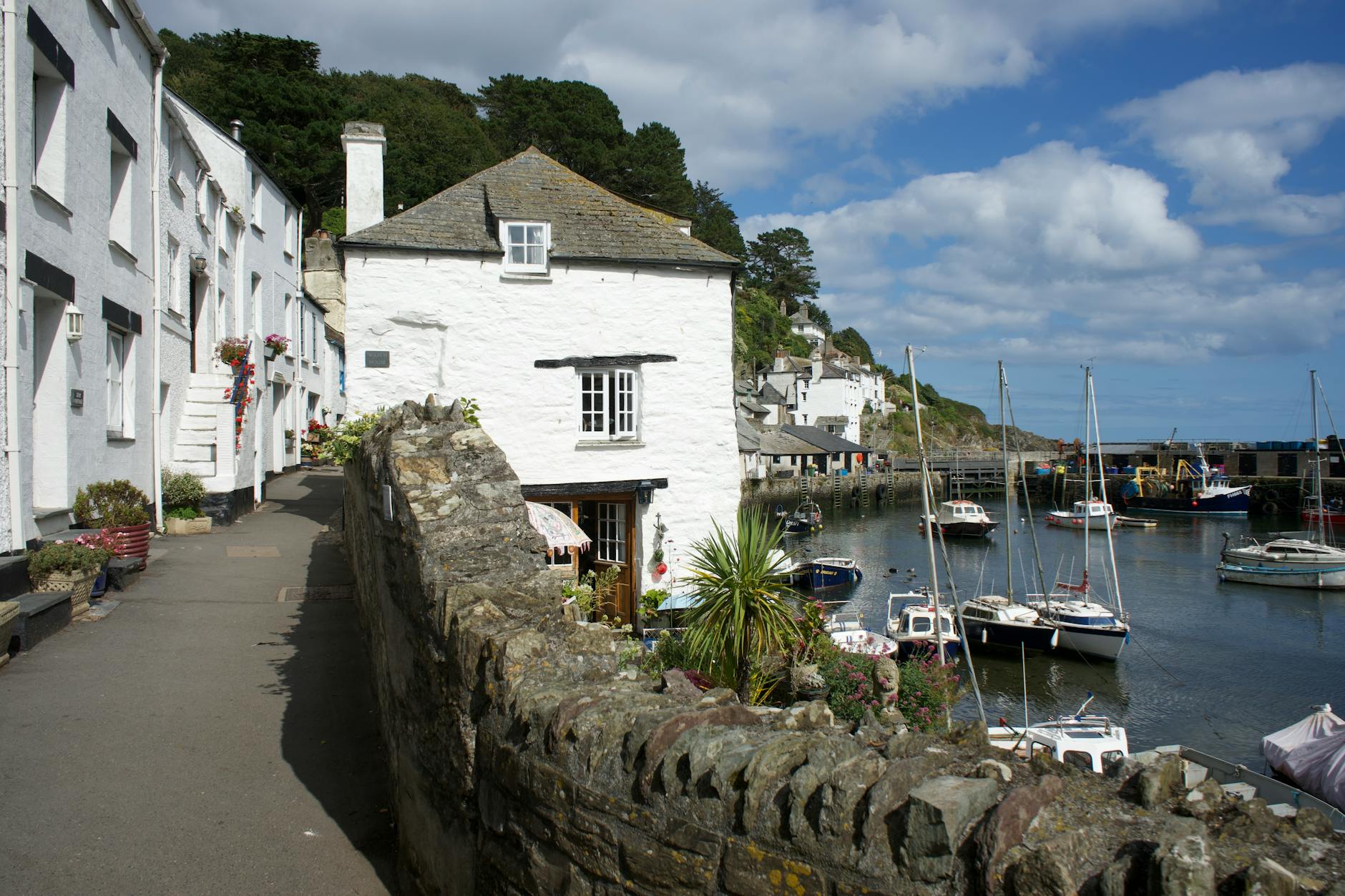 Whitewashed Cornish cottage beside a stone harbour wall with moored yachts and a hillside village under a blue sky.