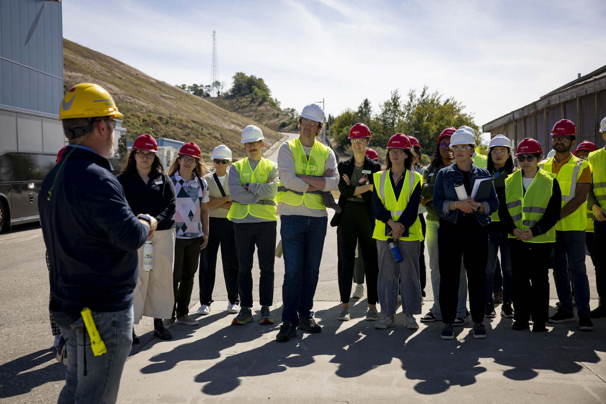 A group of people in safety vests listens to a speaker in a hard hat during an outdoor meeting or briefing.