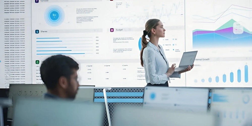 A female professional in a striped shirt holding a laptop and looking at a massive digital wall display filled with data visualizations, charts, and financial analytics in a modern corporate office.