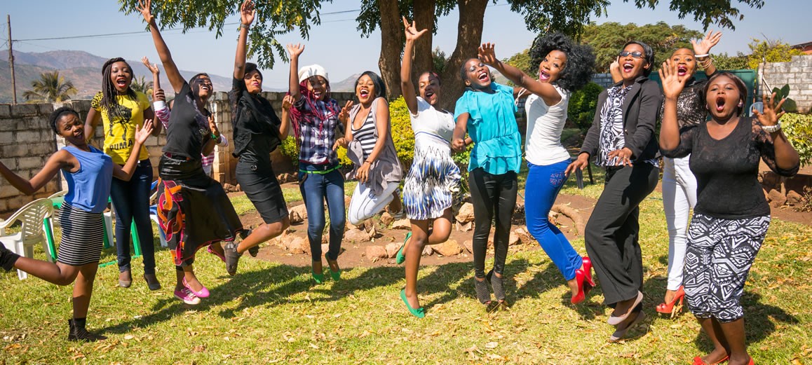 A person wearing headphones sits at a desk with a large curved monitor displaying graphic design software, surrounded by a laptop, smartphone, and potted plant, creating a modern workspace setup.