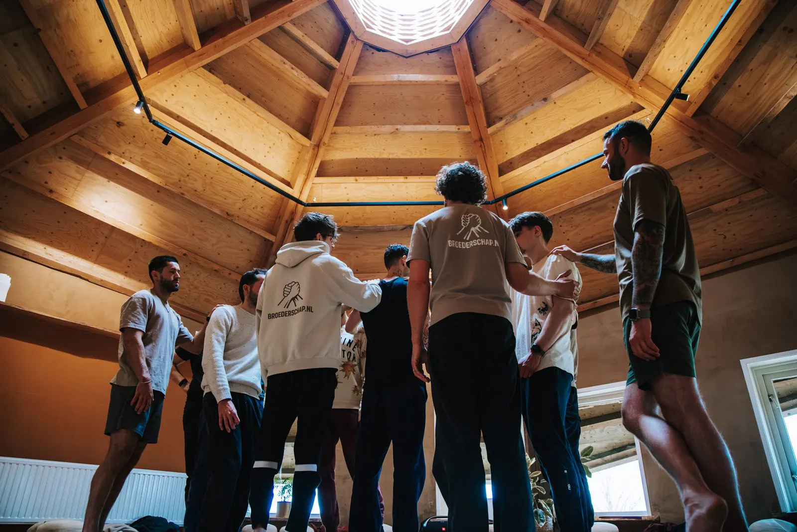 Een groep mannen staat in een kring onder een achthoekig houten plafond, sommigen met een hand op elkaars schouder