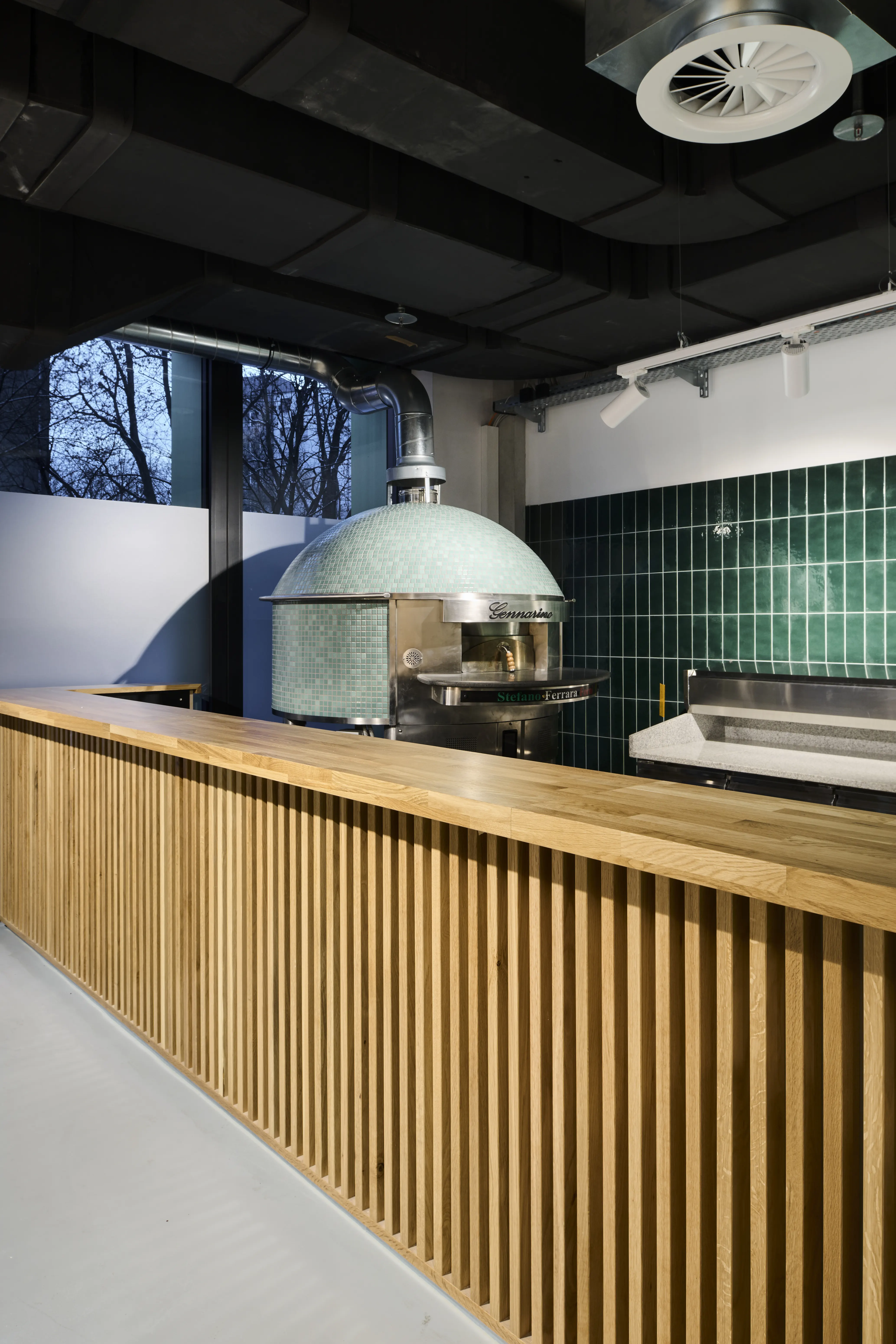 Mint-green tiled dome pizza oven at PARKER BOWLES & FRIENDS behind a wooden slat-fronted counter with oak top and green tiled wall.