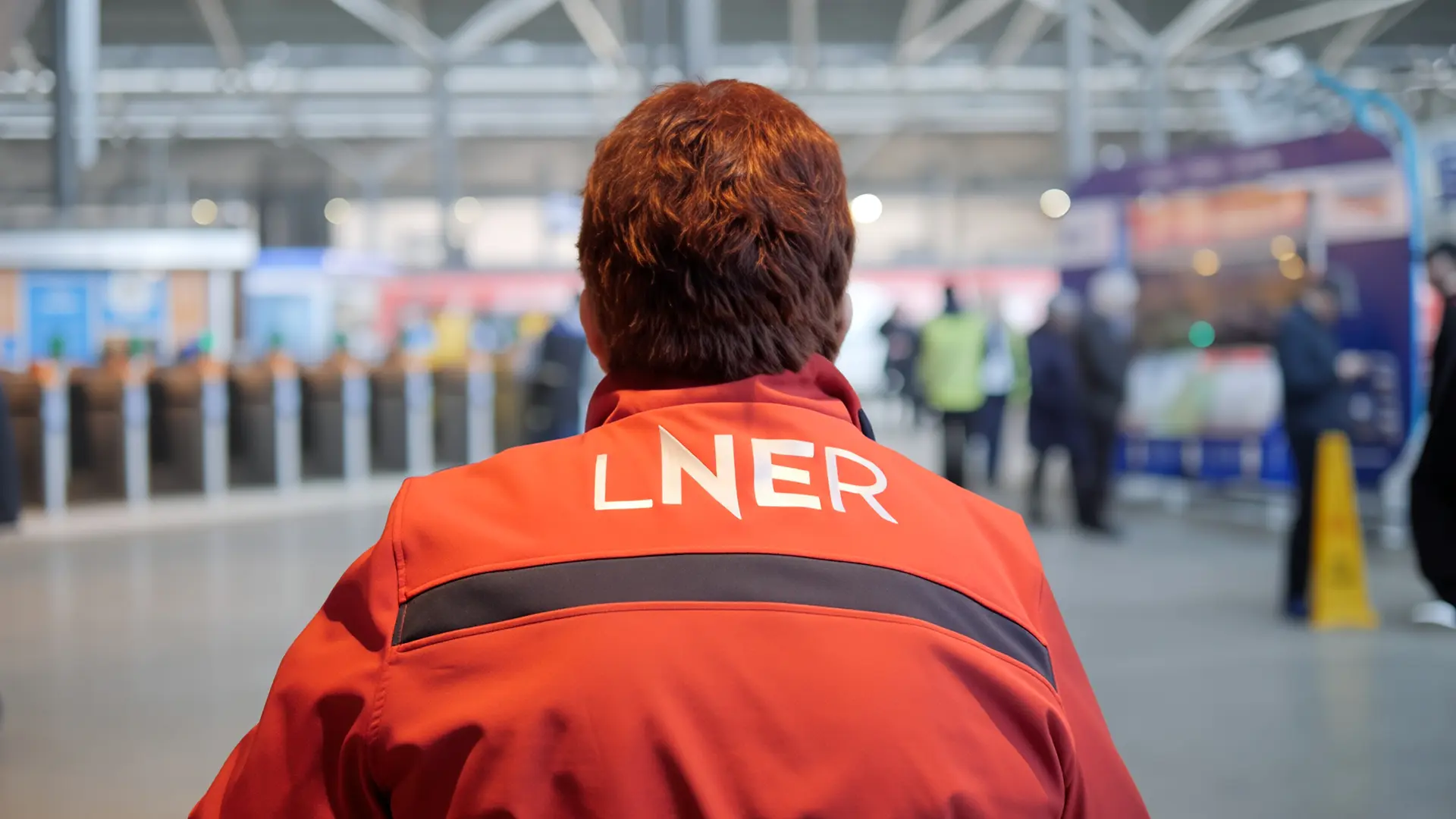 Back view of an LNER colleague wearing a red uniform jacket inside a busy station concourse.