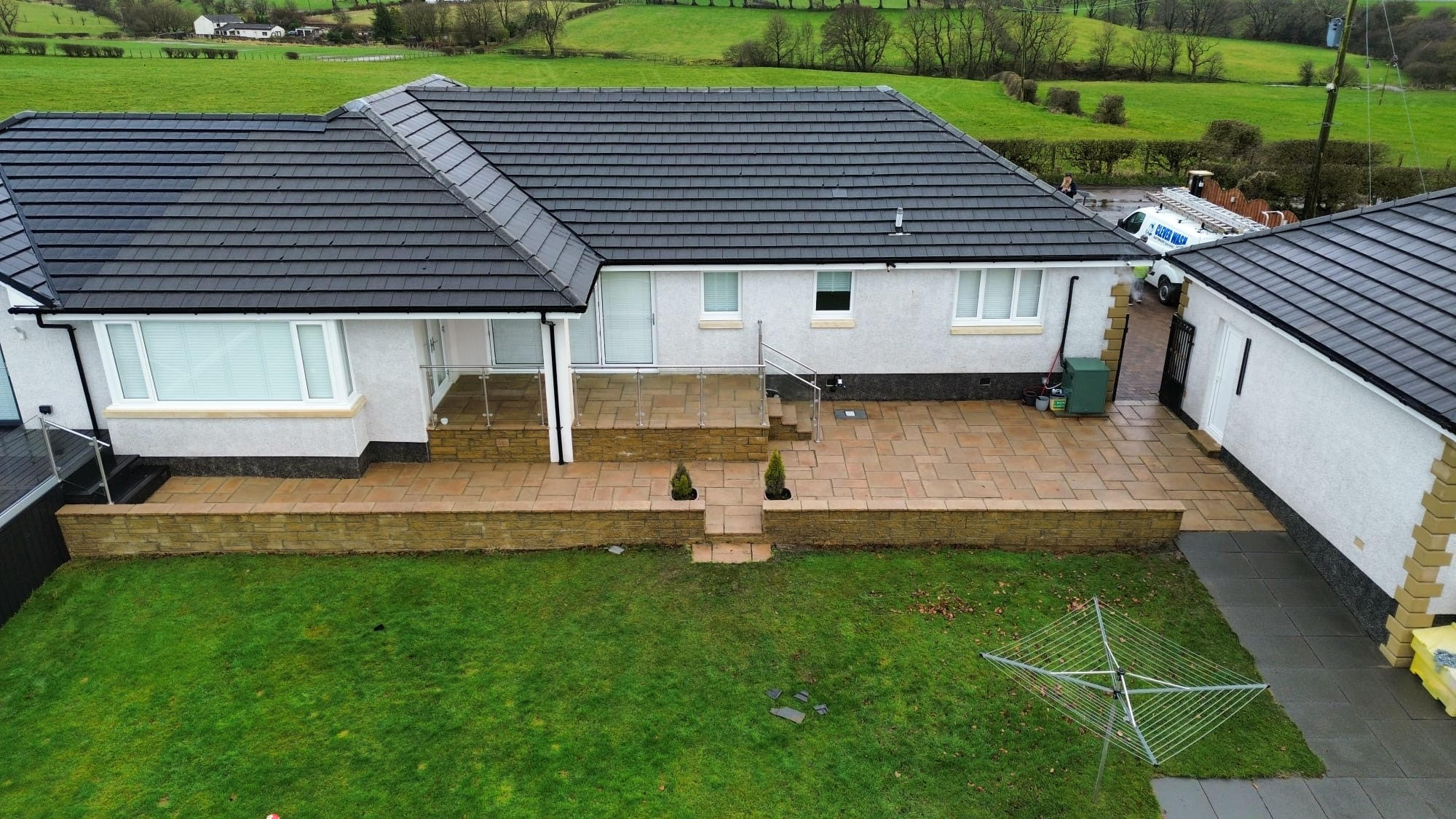 Aerial view of a single-story house with a black tiled roof, white walls, and a spacious stone patio.