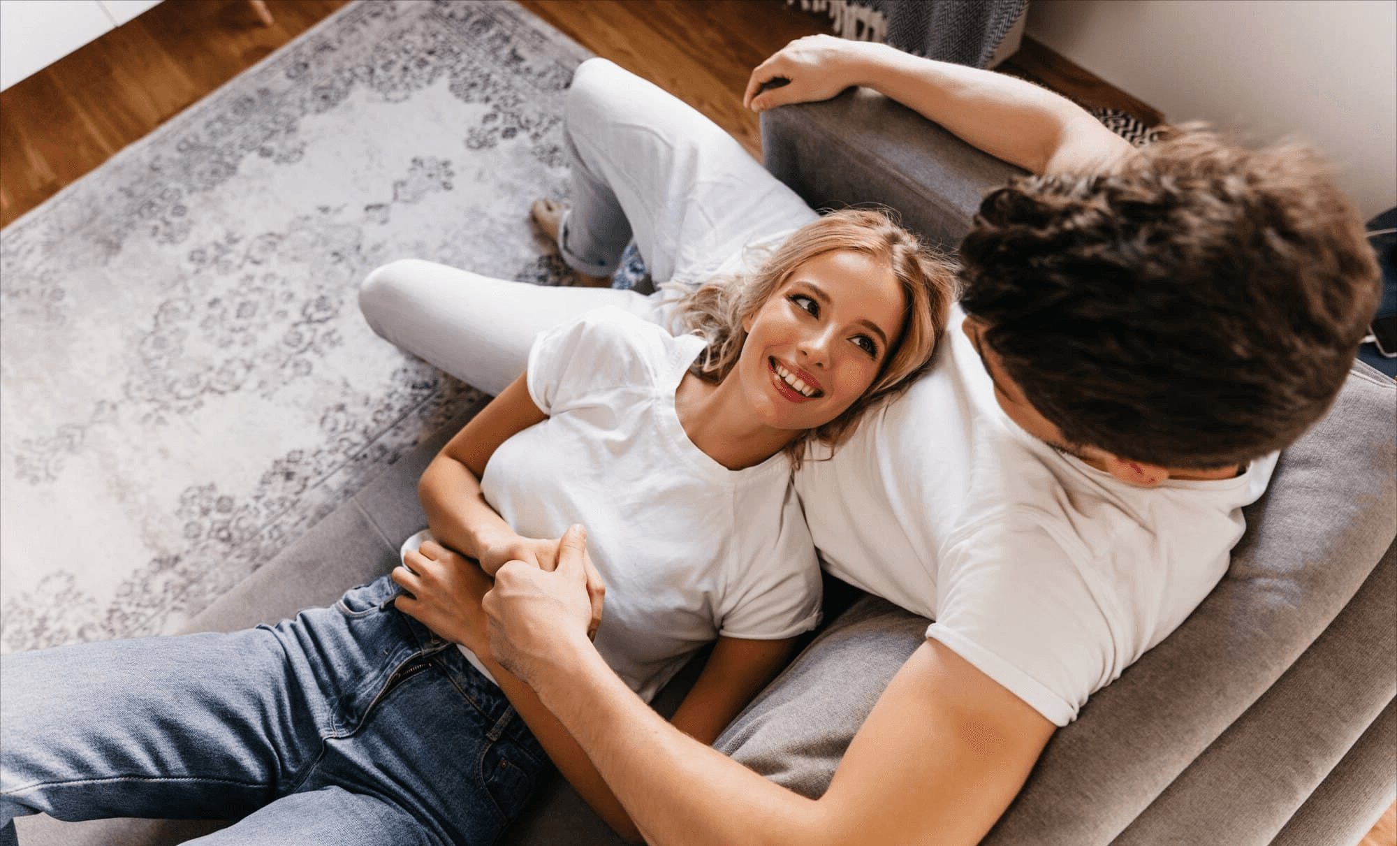 A smiling couple relaxes together on a couch at home, holding hands and enjoying a cozy moment. The bright, welcoming atmosphere reflects the comfort and stability achieved through conventional home loans from Chris Lewis Home Loans.