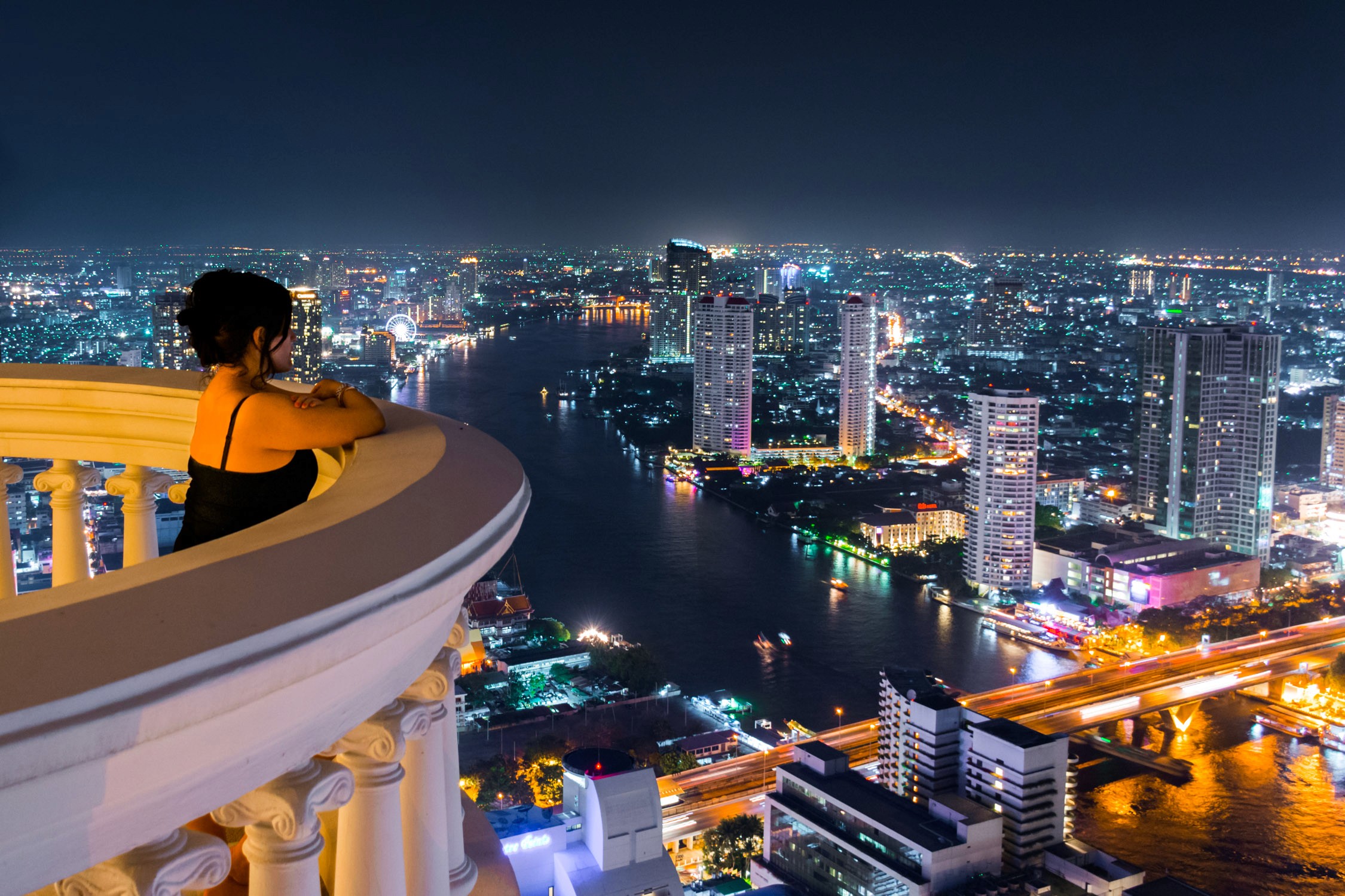 A person in an orange shirt leans on a balcony railing overlooking a brightly lit city skyline and river at night.