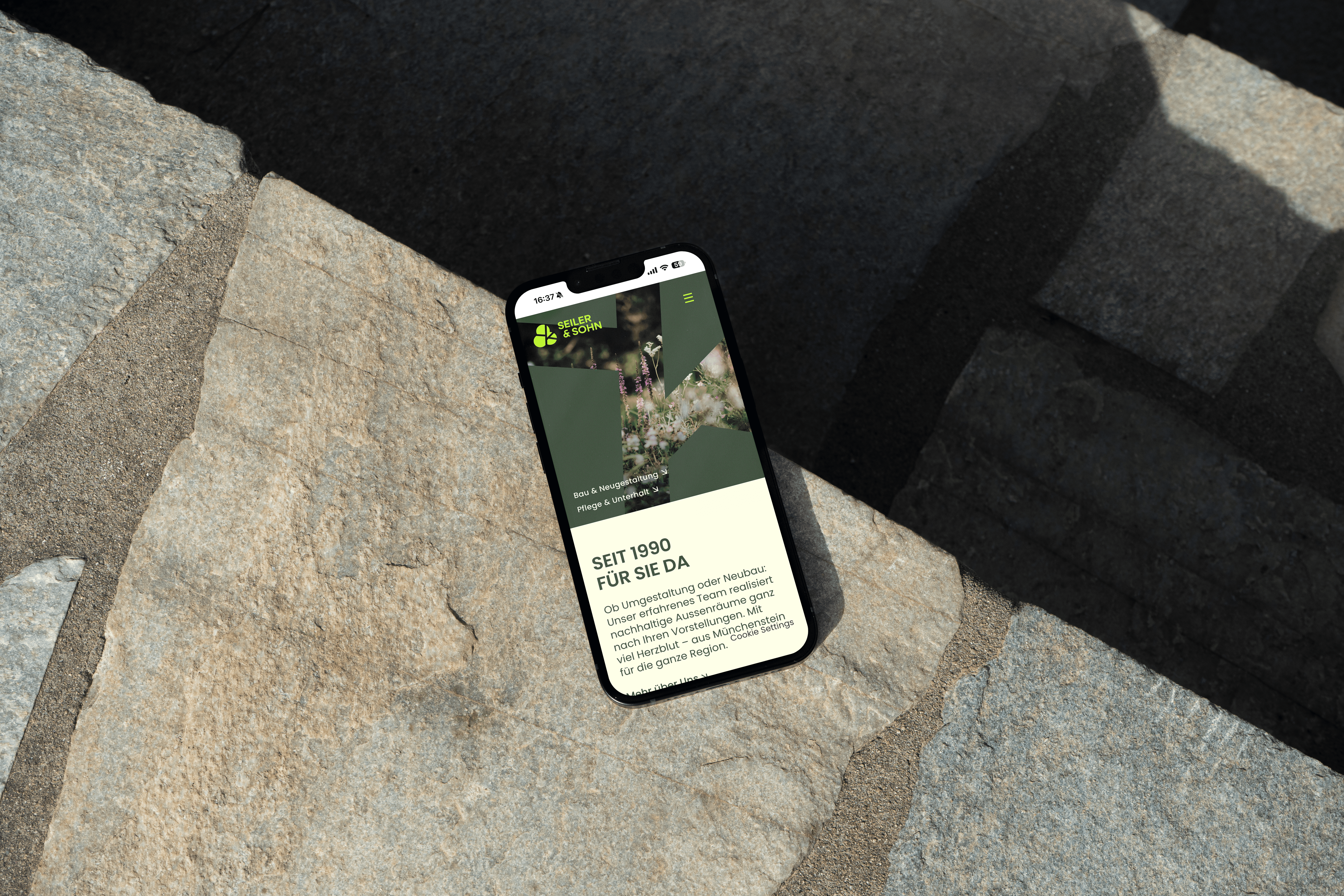 Ein Smartphone liegt schräg auf rauen Steinen und zeigt die mobile Ansicht der Seiler & Sohn Website mit dem Slogan SEIT 1990 FÜR SIE DA und Dienstleistungsübersicht.