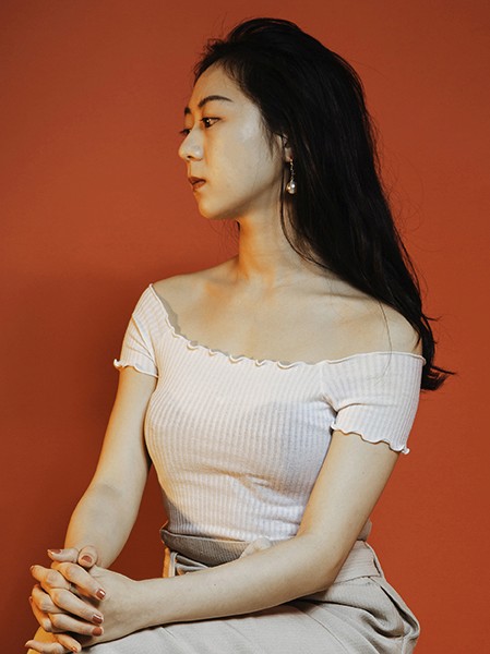 A woman with long, dark hair sits against a solid orange-brown wall. She wears a white, off-the-shoulder, ribbed top with short, ruffled sleeves and a light beige skirt. Her hands are clasped in her lap, and she looks to the left of the frame. She wears small earrings. The lighting is soft and evenly distributed.