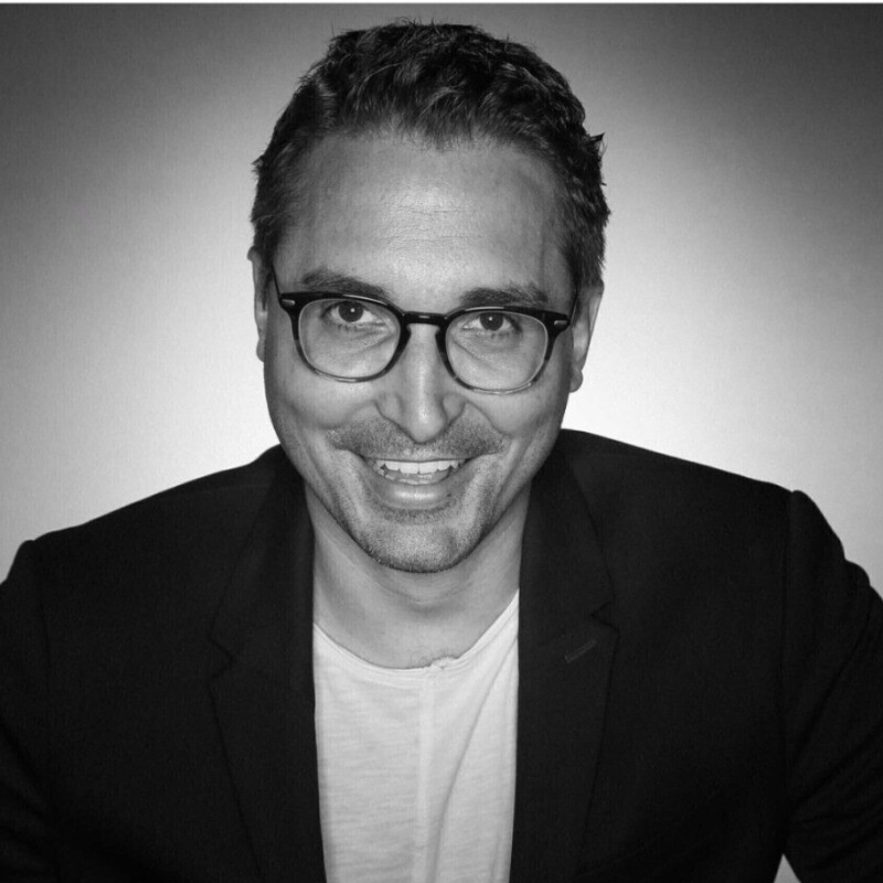 Mann in beigem Hemd und Brille sitzt auf Sofa und blickt in die Kamera