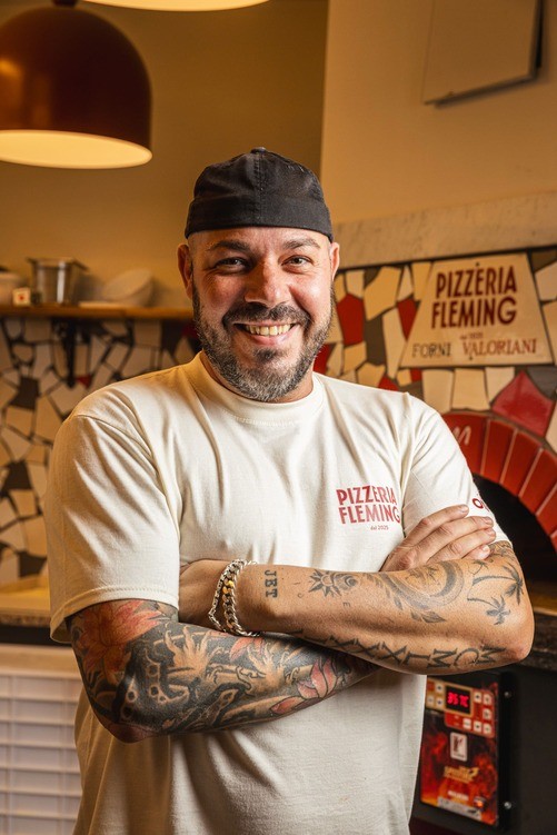 A smiling chef stands confidently with arms crossed in a vibrant kitchen setting, showcasing a welcoming atmosphere.