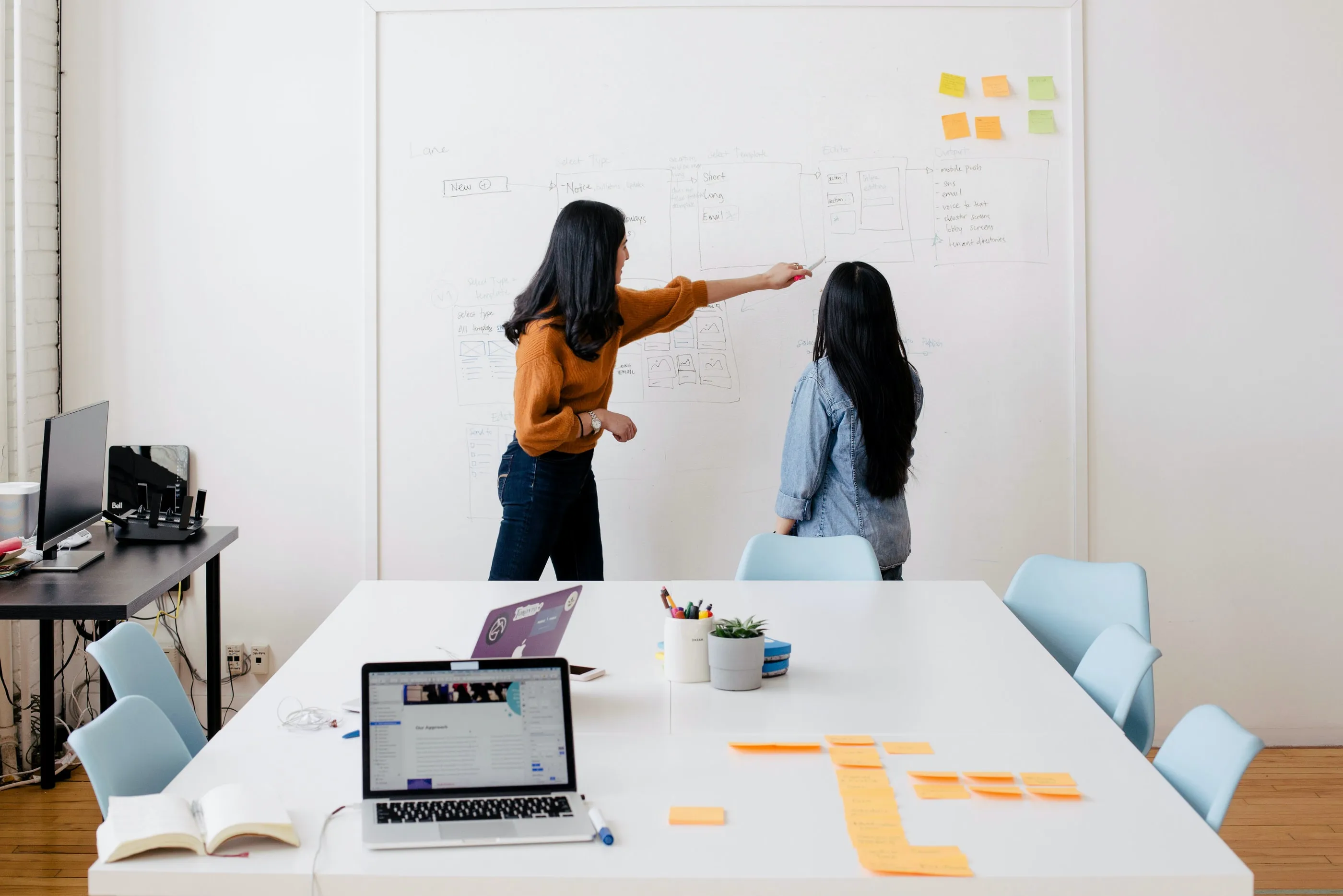 Two women brainstorming and discussing ideas in front of a whiteboard in a modern office setup