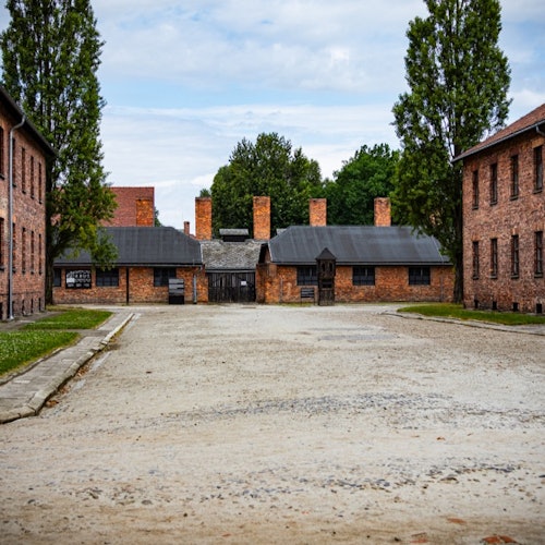 Een ruime, lege binnenplaats wordt geflankeerd door rode bakstenen gebouwen met hoge schoorstenen en groen op de achtergrond onder een bewolkte hemel.