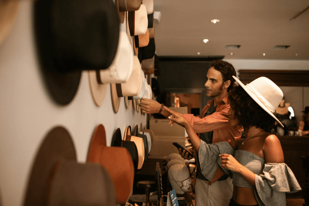 Two people in a shop choosing from a wall of different hats, a literal visual metaphor for how instructional designers must 'wear many hats' and adapt to diverse job roles.