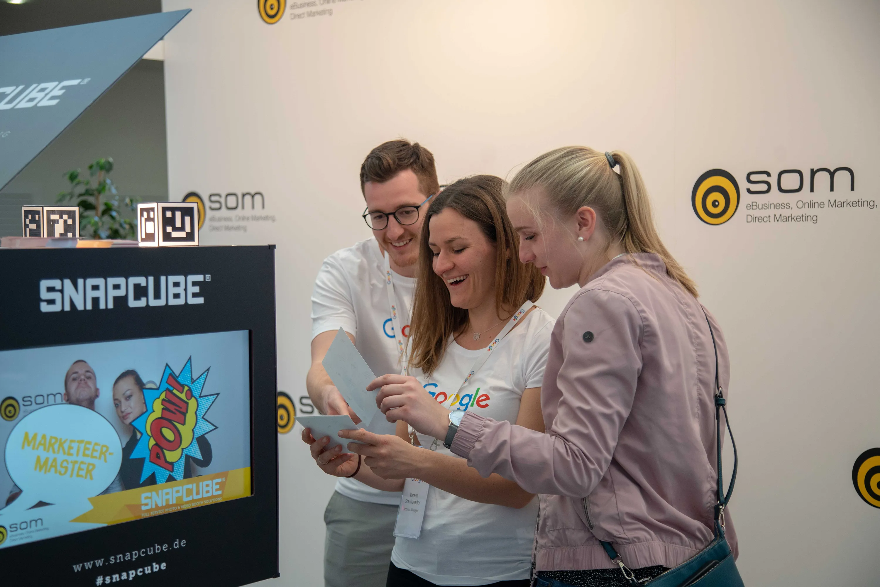 Bei der SOM Messe sichten Mitarbeiter von Google ihre Fotos. Das Gehäuse der Fotobox zeigt den Sponsor SnapCube®. Die Bildschirme zeigen eine Slideshow der aufgenommenen Fotos.