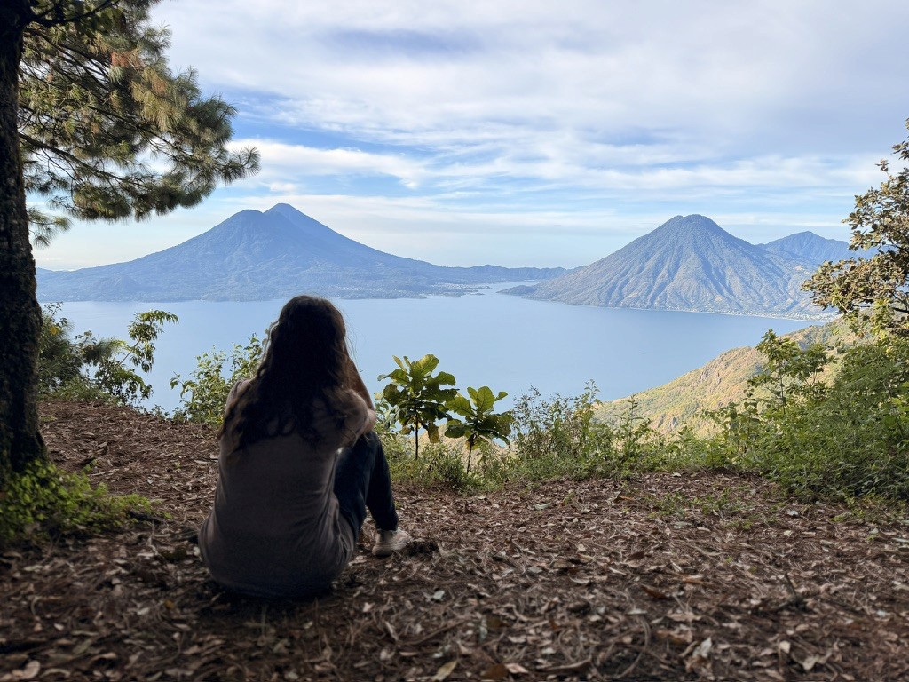 A person seated beneath trees overlooking a lake and surrounding mountains.