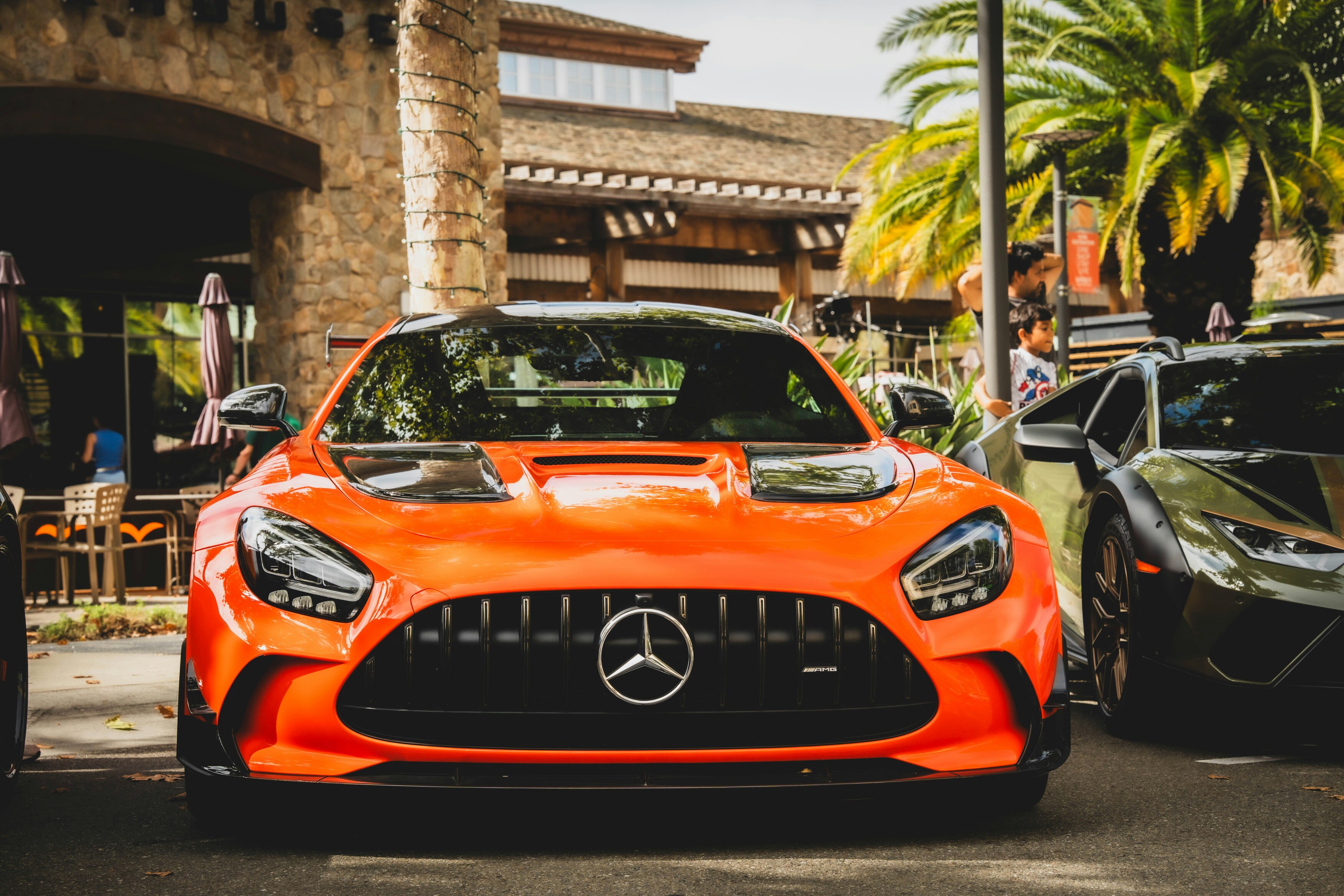 A vibrant orange Mercedes sports car is parked outdoors, with sleek black accents and a distinctive front grille, surrounded by tropical palm trees and a stone building in the background.